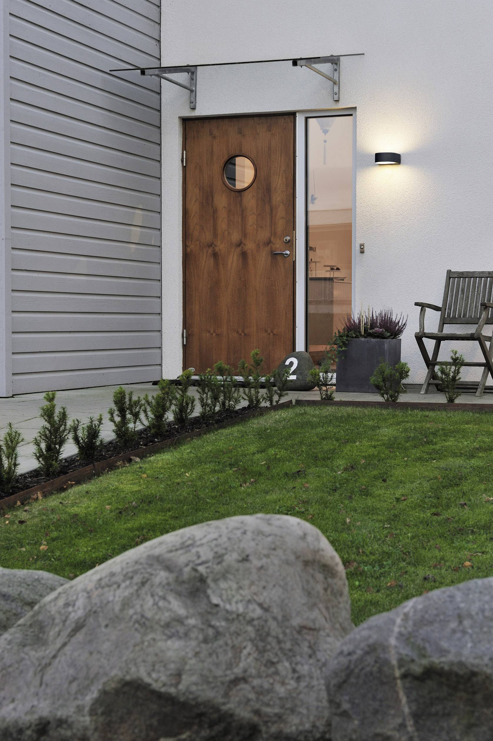 Holztür mit rundem Fenster an modernem Haus, beleuchtet von Außenlampe. Kleiner Garten mit Rasen und Steinen im Vordergrund.