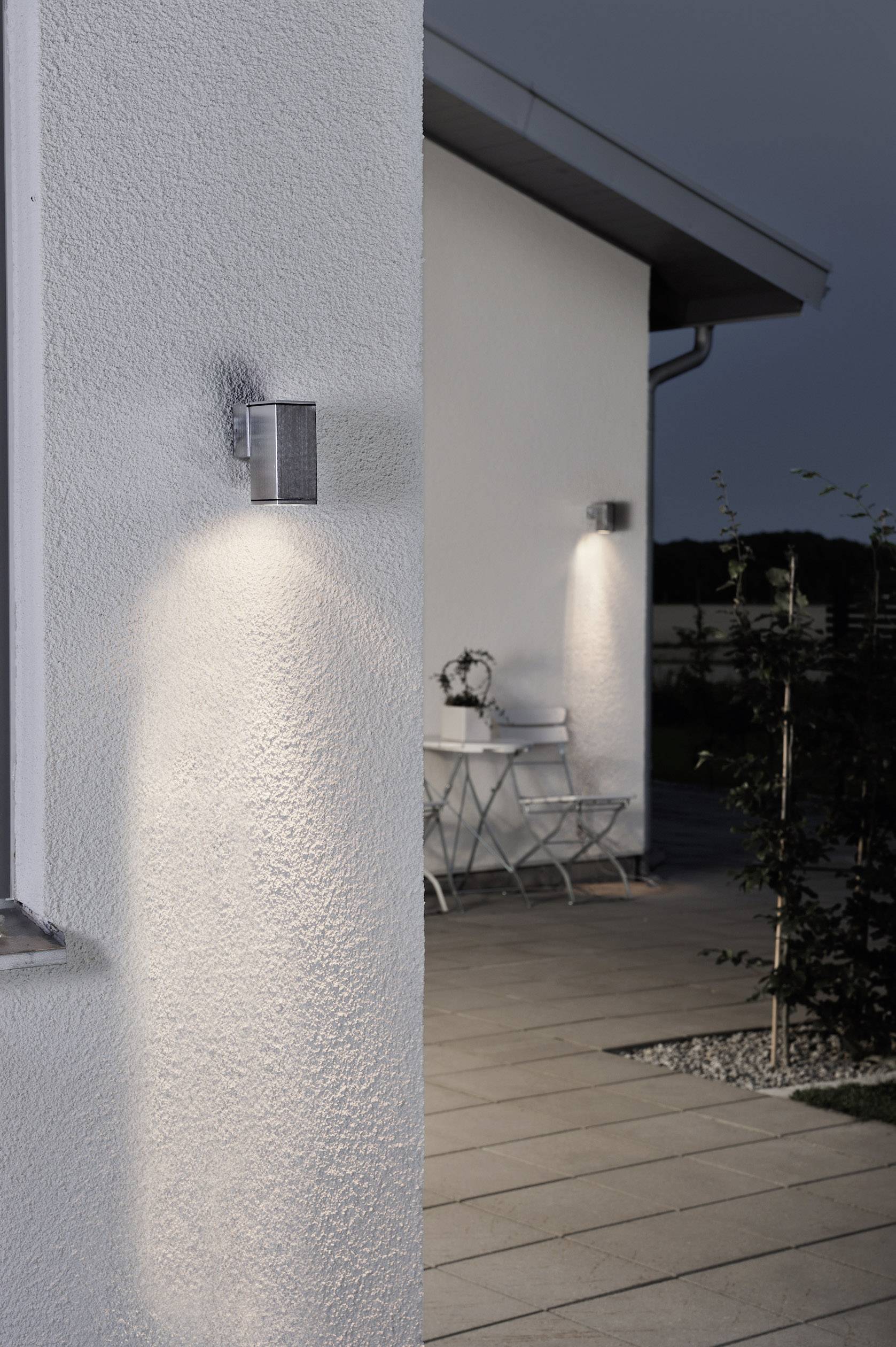 Außenansicht eines Hauses bei Dämmerung mit eingeschalteten Wandlampen, die warmes Licht auf eine Terrasse mit Tisch und Stühlen werfen.