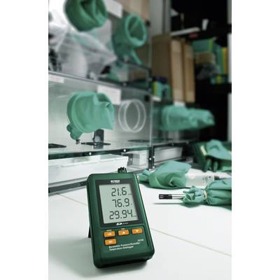 Ein digitales Thermo-Hygrometer zeigt Temperaturen auf einem Tisch an. Im Hintergrund sind Laborgeräte und grüne Schutzhandschuhe zu sehen.