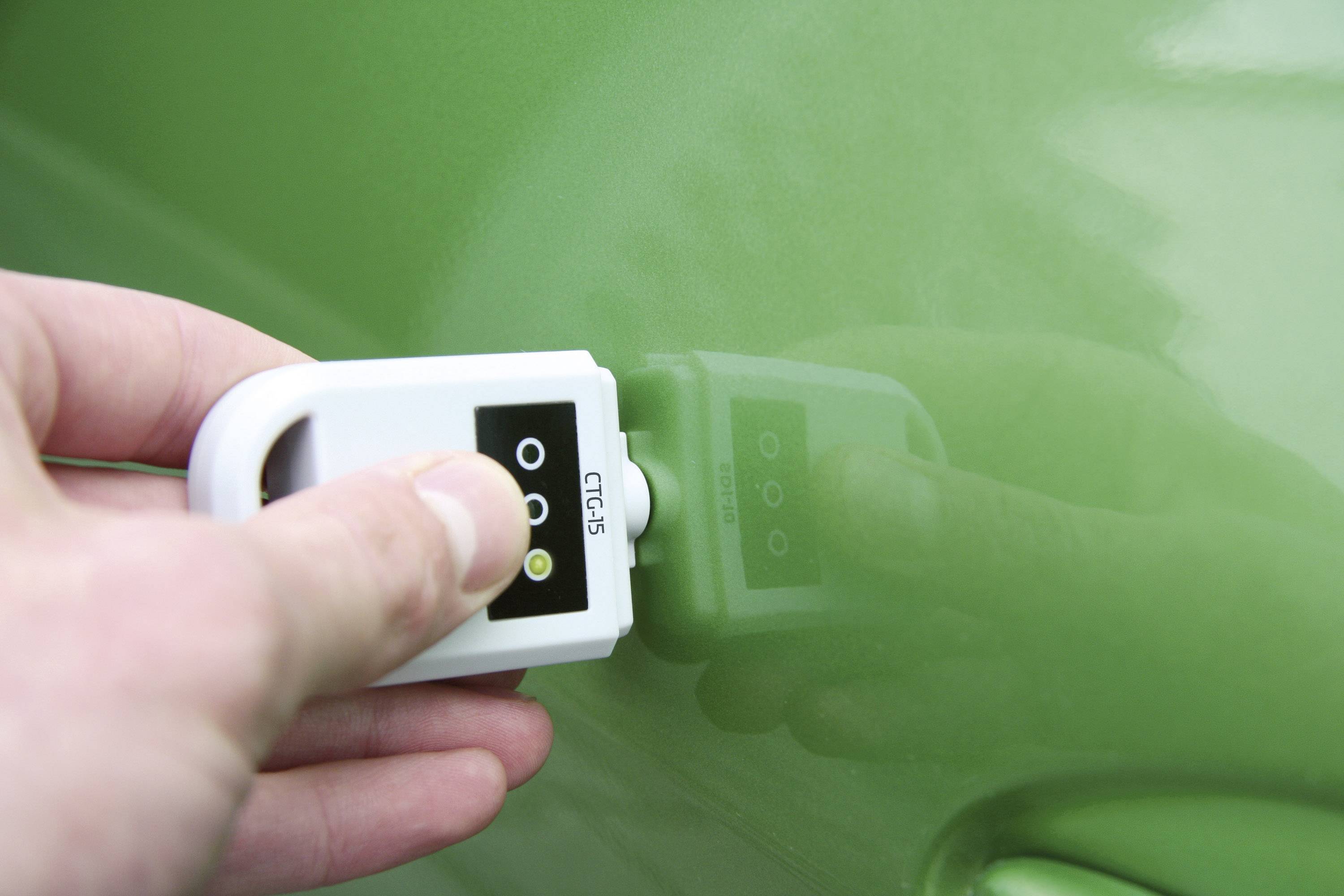 Eine Hand hält ein Gerät gegen ein grünes Auto, um die Lackdicke zu messen. Das Display zeigt Werte an.