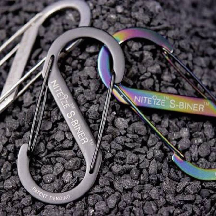 Zwei S-Biner von Nite Ize, einer metallisch grau, der andere schillernd bunt, liegen auf schwarzem Untergrund.