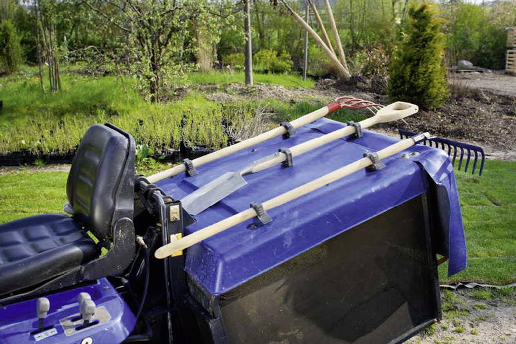 Rasenmähertraktor mit angehängten Gartenwerkzeugen wie Schaufel, Harke und Spaten. Im Hintergrund ein Garten mit Bäumen und Sträuchern.