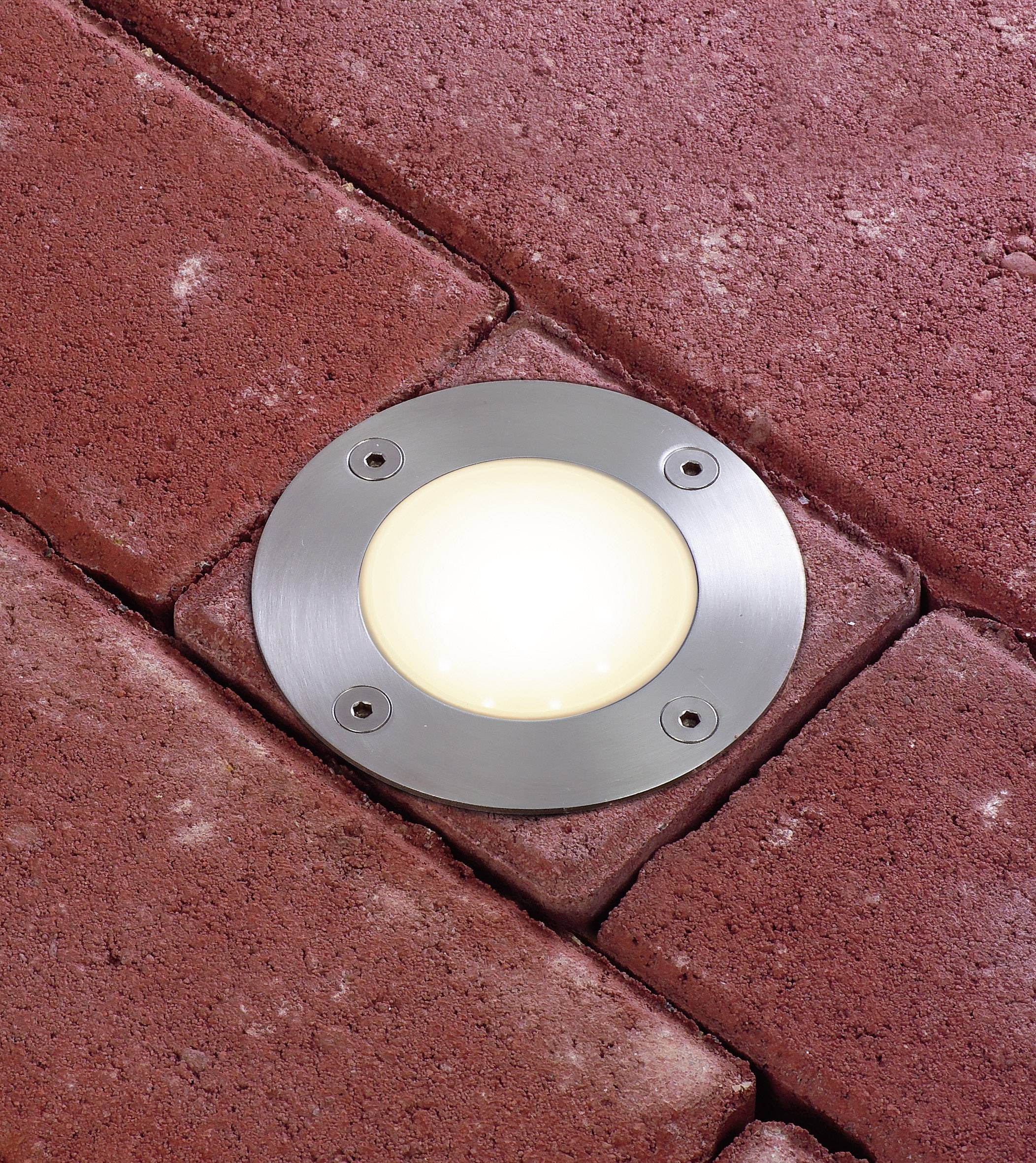 Eine runde Bodenleuchte ist in rotem Pflasterstein eingebaut und leuchtet hell.