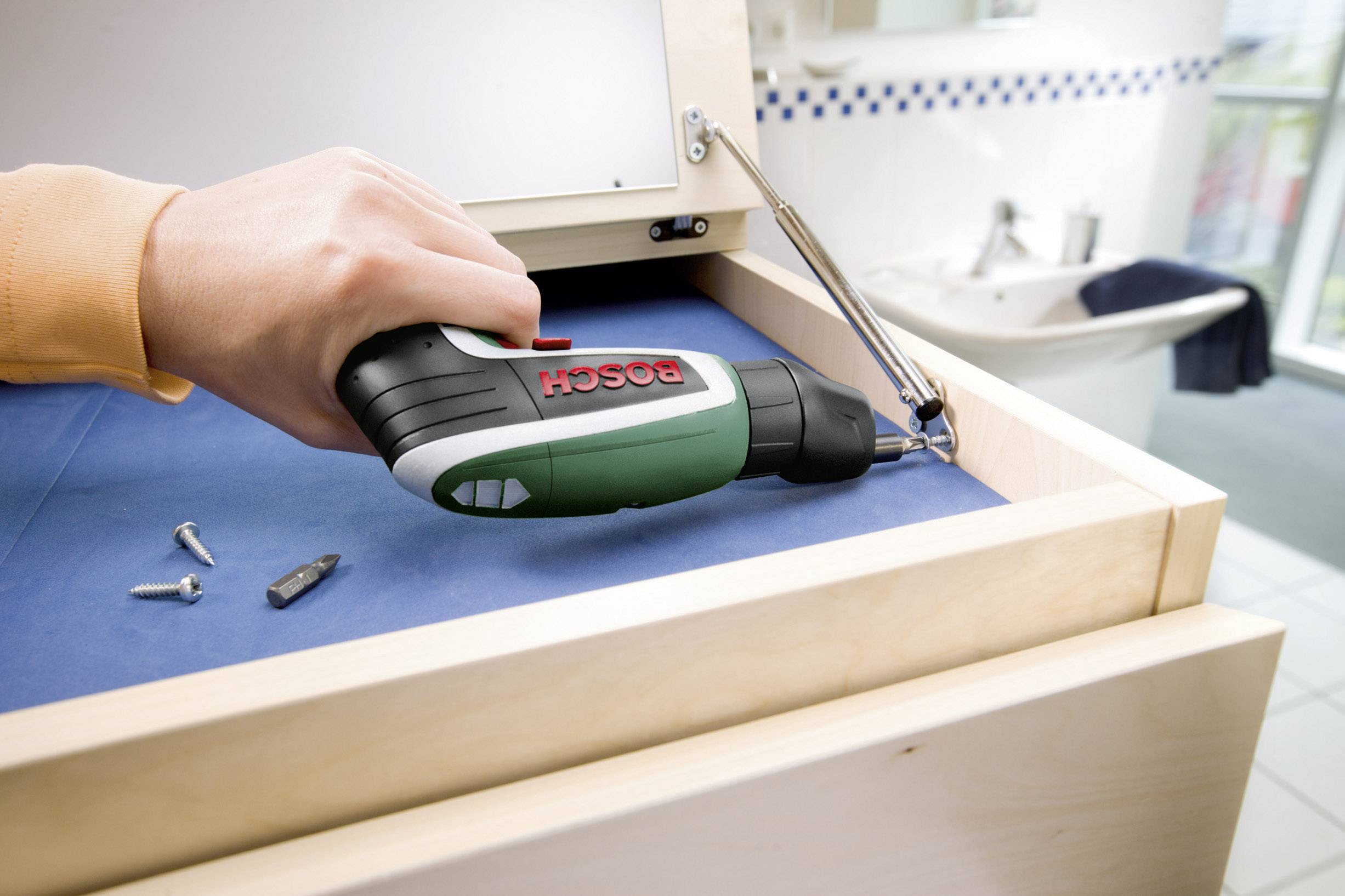 Eine Person verwendet einen Akkuschrauber, um eine Schraube in einem Holzschrank festzuziehen. Im Hintergrund ist ein Waschbecken.