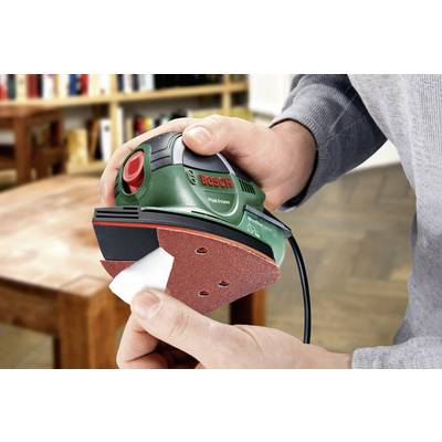 Eine Person hält einen grünen Bosch-Multischleifer und wechselt das Schleifpapier. Im Hintergrund ist ein Holztisch sichtbar.