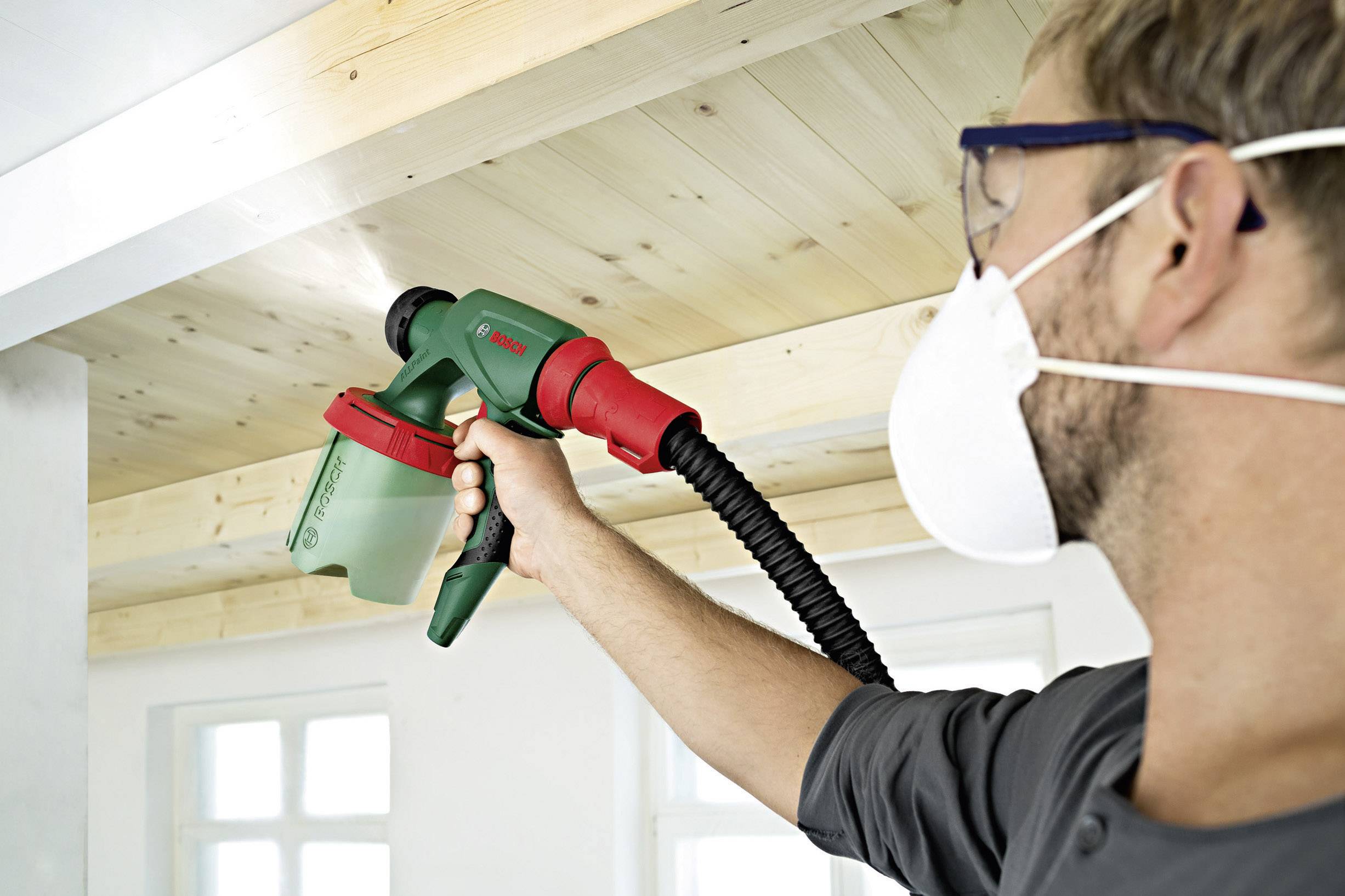 Ein Mann sprüht Farbe an eine Holzdecke mit einer Farbspritzpistole. Er trägt eine Schutzbrille und eine Atemschutzmaske.