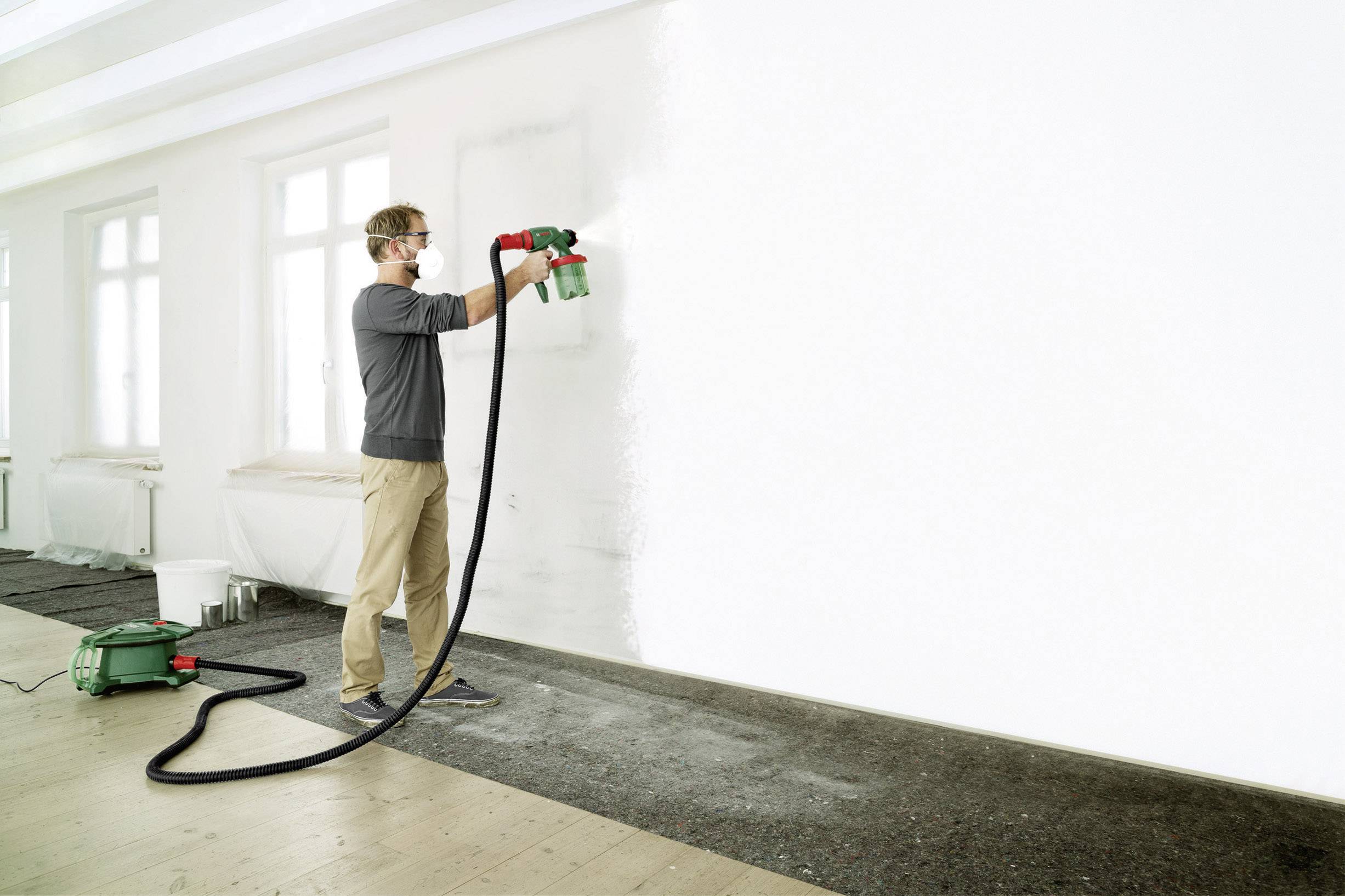 Ein Mann sprüht weiße Farbe mit einem Sprühgerät an eine große, leere Wand in einem hell erleuchteten Raum.