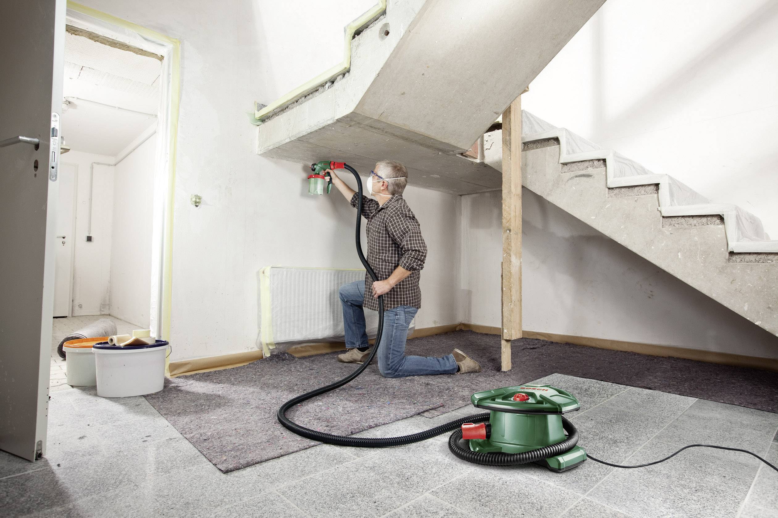 Ein Mann kniet auf dem Boden und malt mit einem Sprühgerät die Wand unter einer Treppe. Ein Eimer und ein Sauggerät sind zu sehen.
