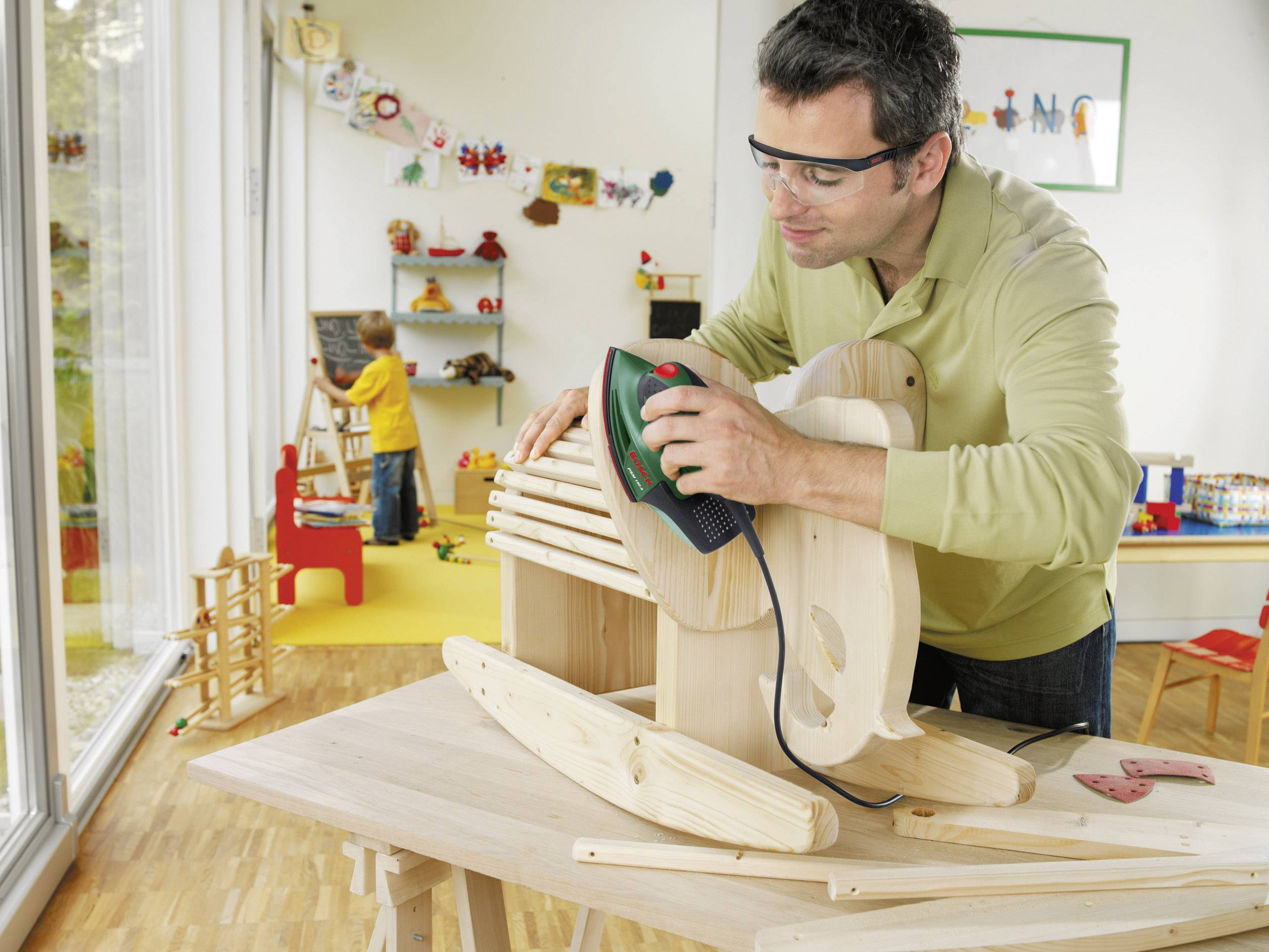 Mann schleift ein Holzpferd mit einem Elektrowerkzeug. Im Hintergrund spielt ein Kind in einem bunt dekorierten Raum.