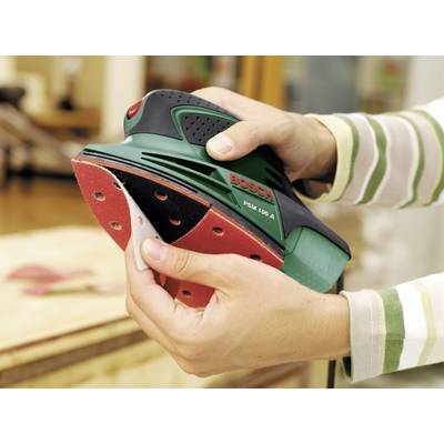 Person hält einen grünen 'Bosch PSM 100 A'-Schleifer und wechselt Schleifpapier. Im Hintergrund ein unscharfer Holztisch.