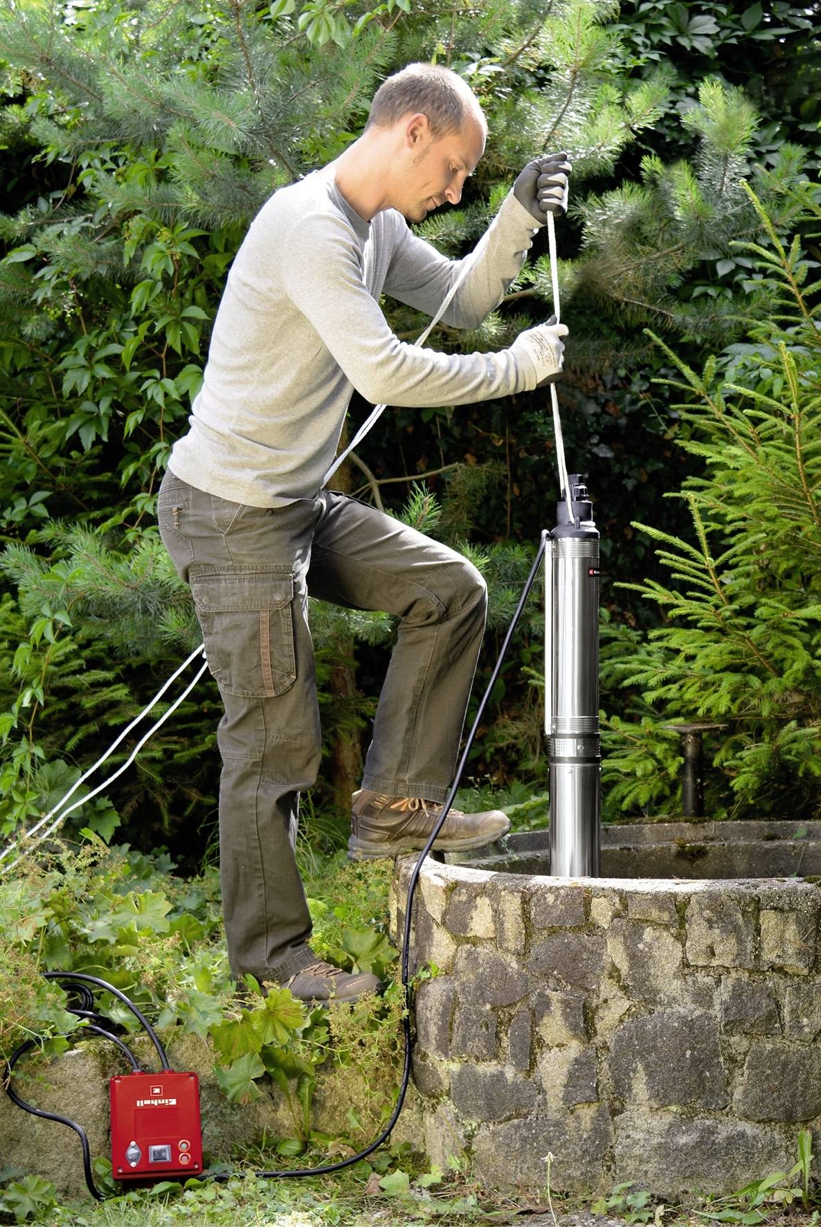 Eine Person installiert eine Pumpe in einem Brunnen, umgeben von dichter Vegetation. Sie benutzt eine Handschuh, um das Gerät zu sichern.