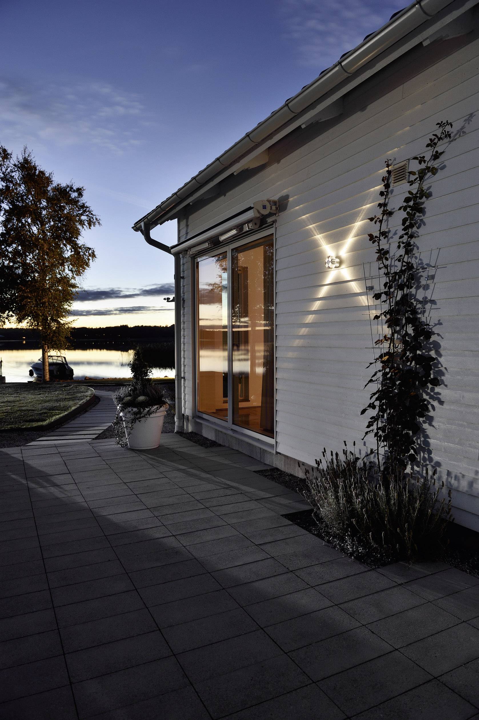 Ein modernes Haus mit Holzfassade bei Sonnenuntergang, Fenster spiegelt das Abendlicht. Ein Baum und ein ruhiger See im Hintergrund.