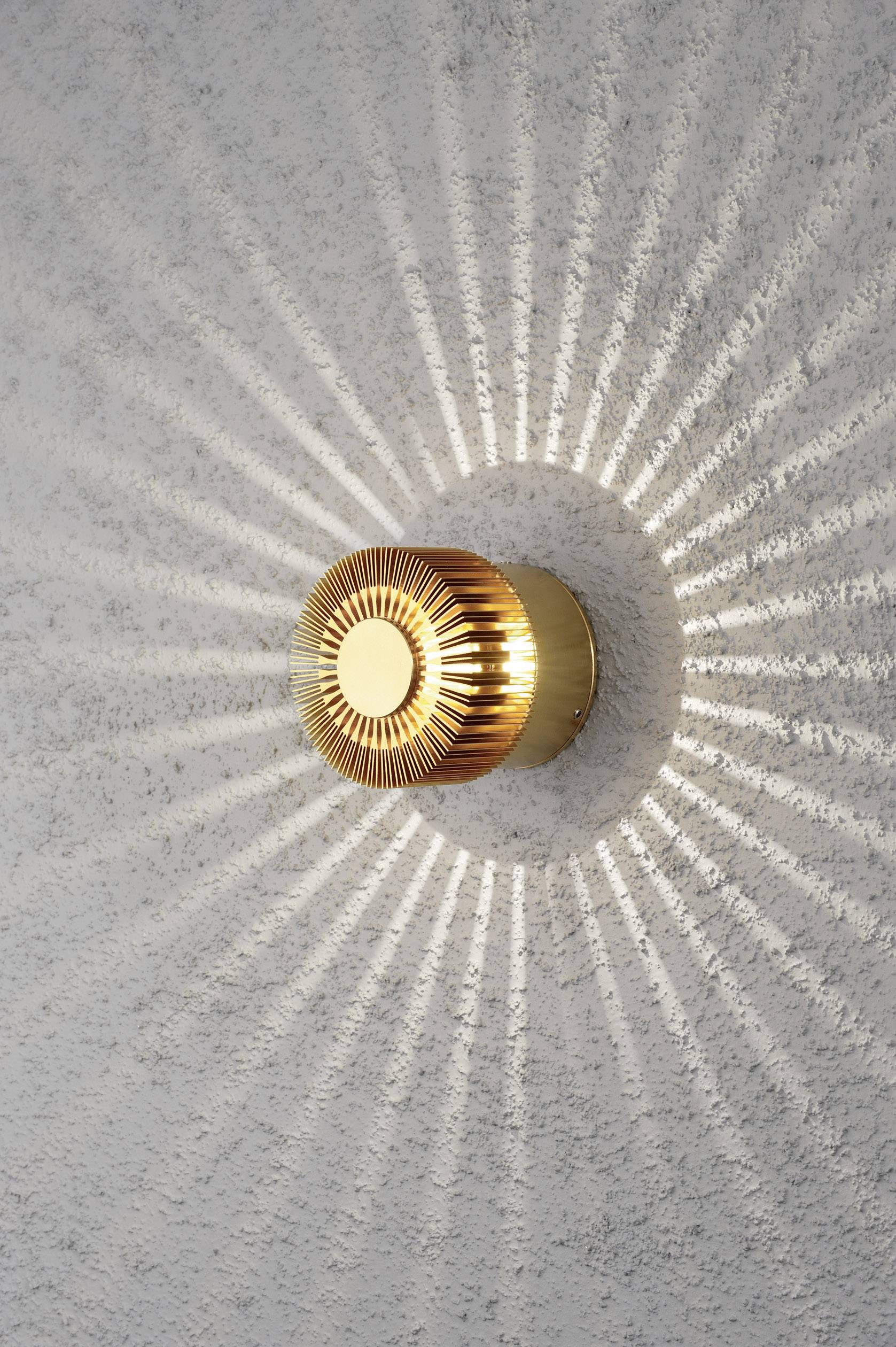 Wandleuchte mit goldenem Design strahlt kreisförmige Lichtmuster an graue Wand, schafft dekorativen Lichteffekt.