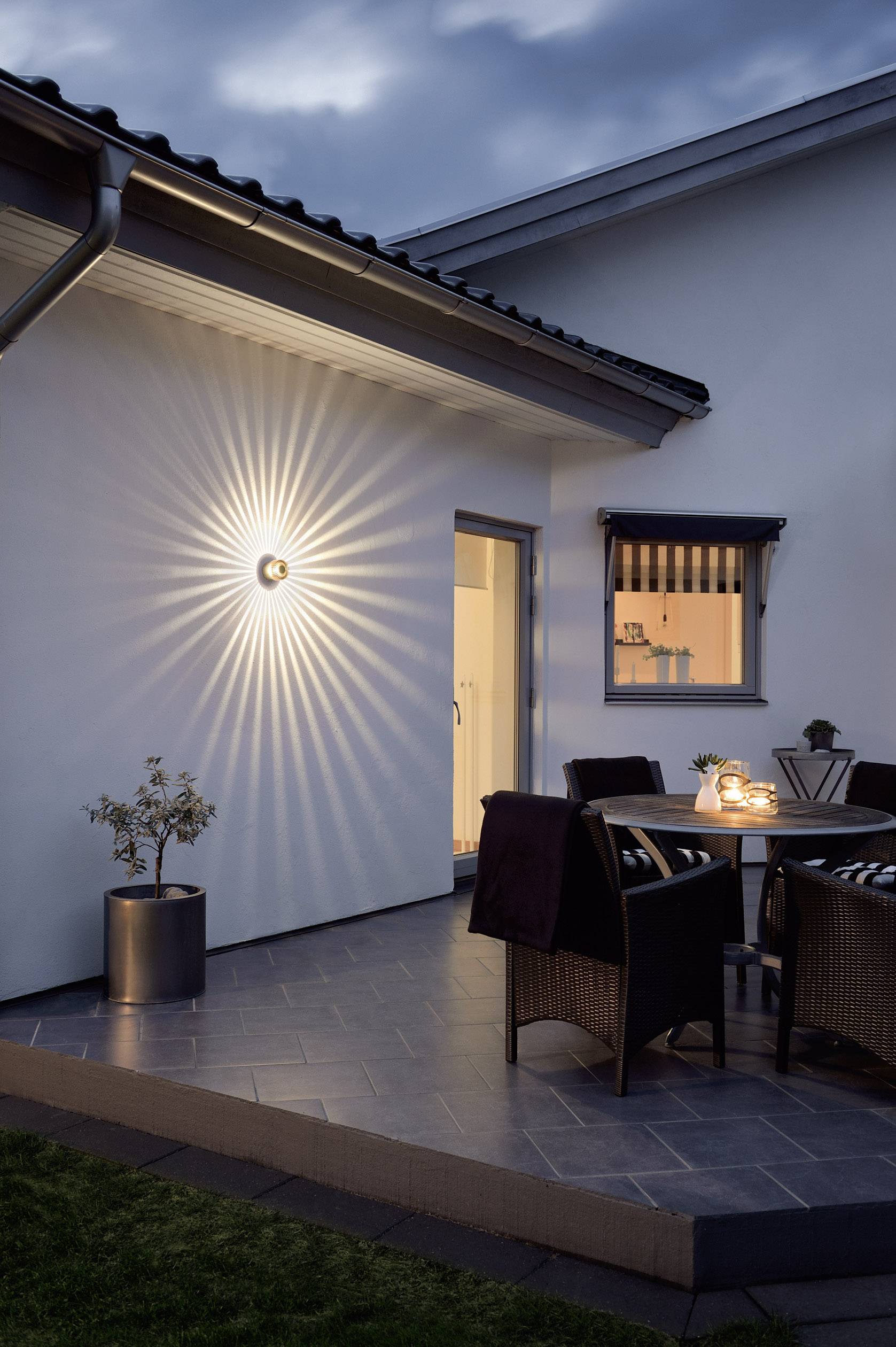 Außenansicht einer Terrasse mit Tisch und Stühlen bei Dämmerung. Eine Lampe an der Hauswand sorgt für stimmungsvolle Beleuchtung.