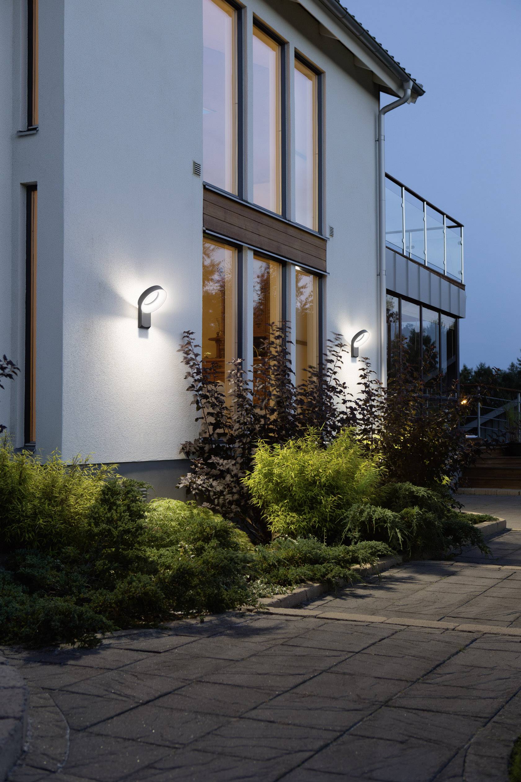 Moderne Hausfassade bei Dämmerung, beleuchtet durch Wandleuchten. Glasschiebetüren und Pflanzen im Vordergrund.
