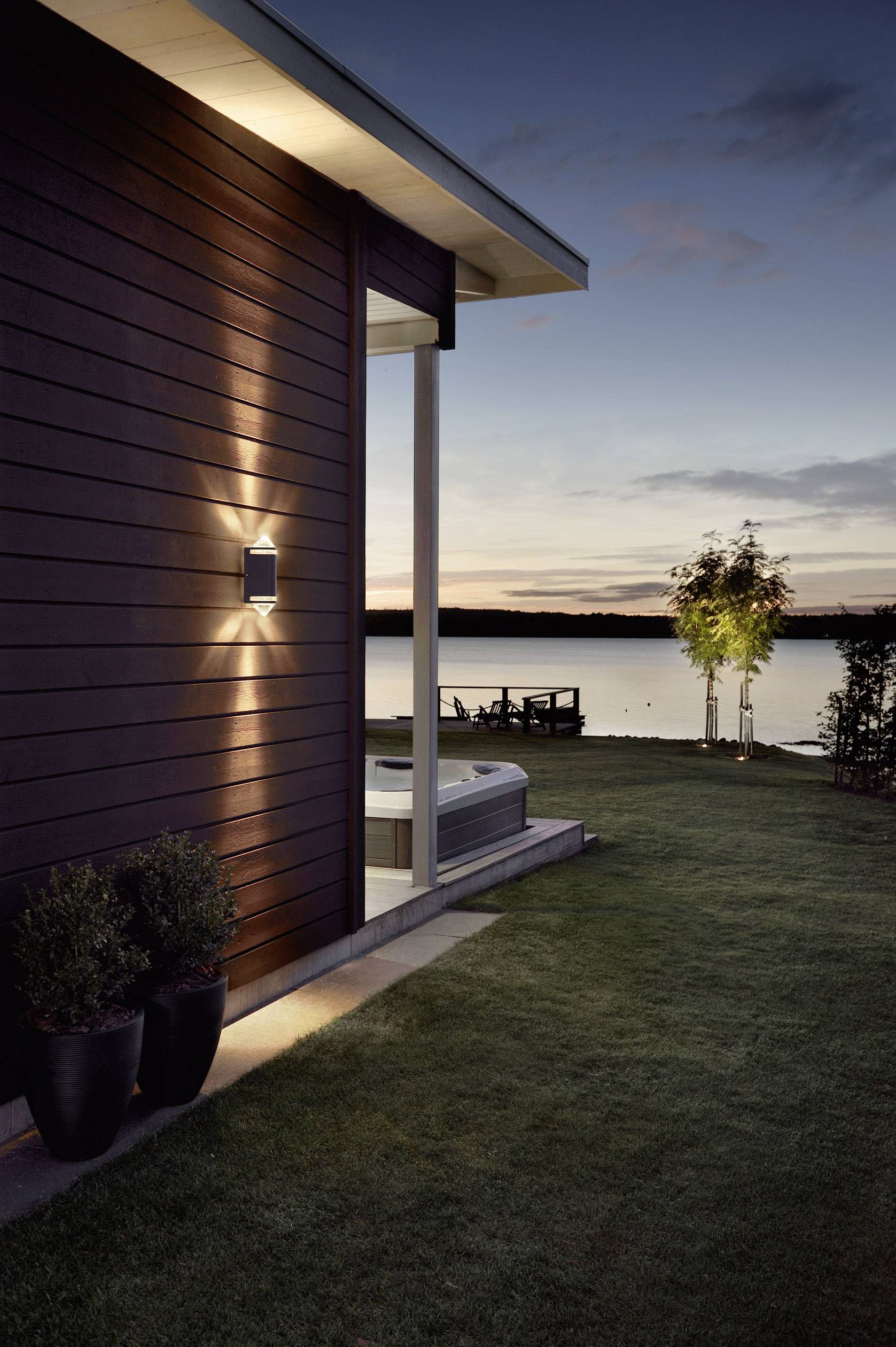 Ein modernes Haus mit Holzfassade bei Dämmerung, Blick auf einen See. Eine Terrasse mit Jacuzzi und stimmungsvoller Außenbeleuchtung.