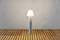 Eine moderne Stehlampe mit konischem Lampenschirm steht auf einem hölzernen Boden vor einer grauen Wand.