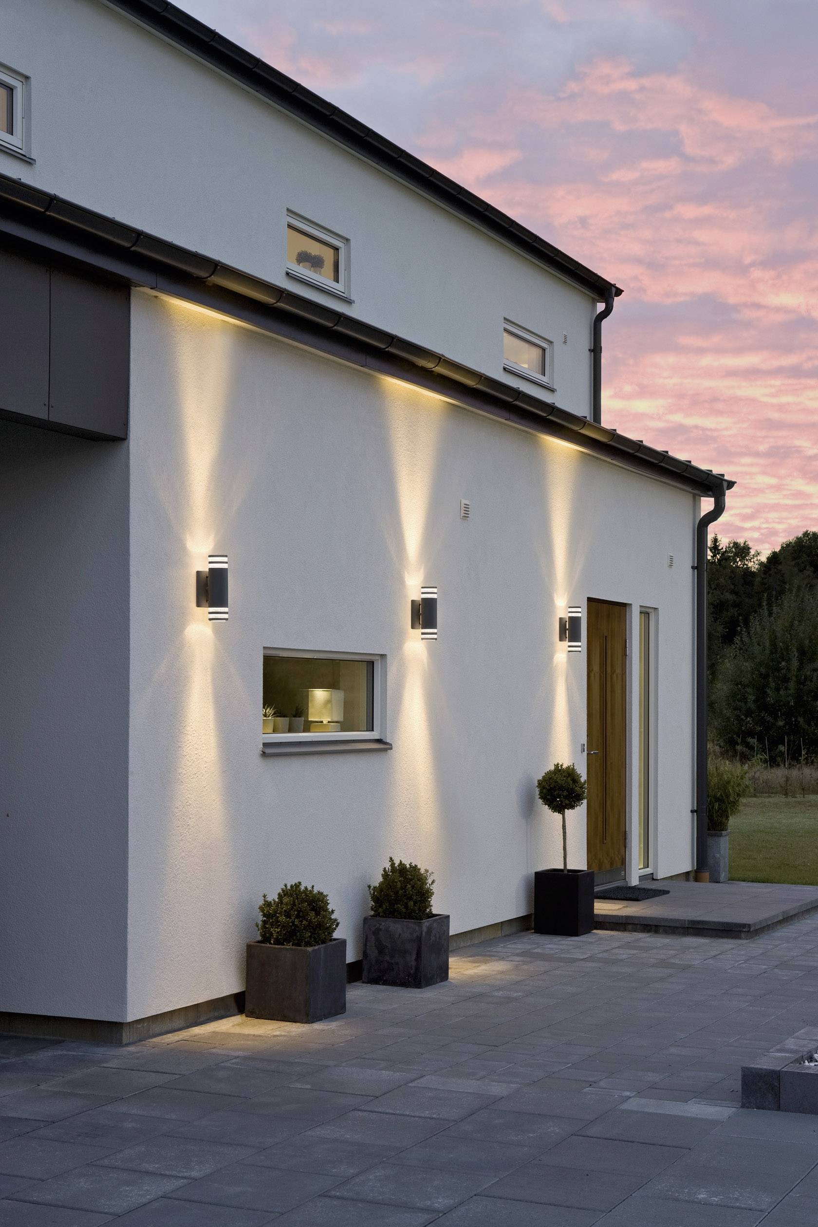 Modernes weißes Haus bei Sonnenuntergang. Außenbeleuchtung hebt geometrisches Design hervor. Einfache Pflanzen schmücken den Eingang.