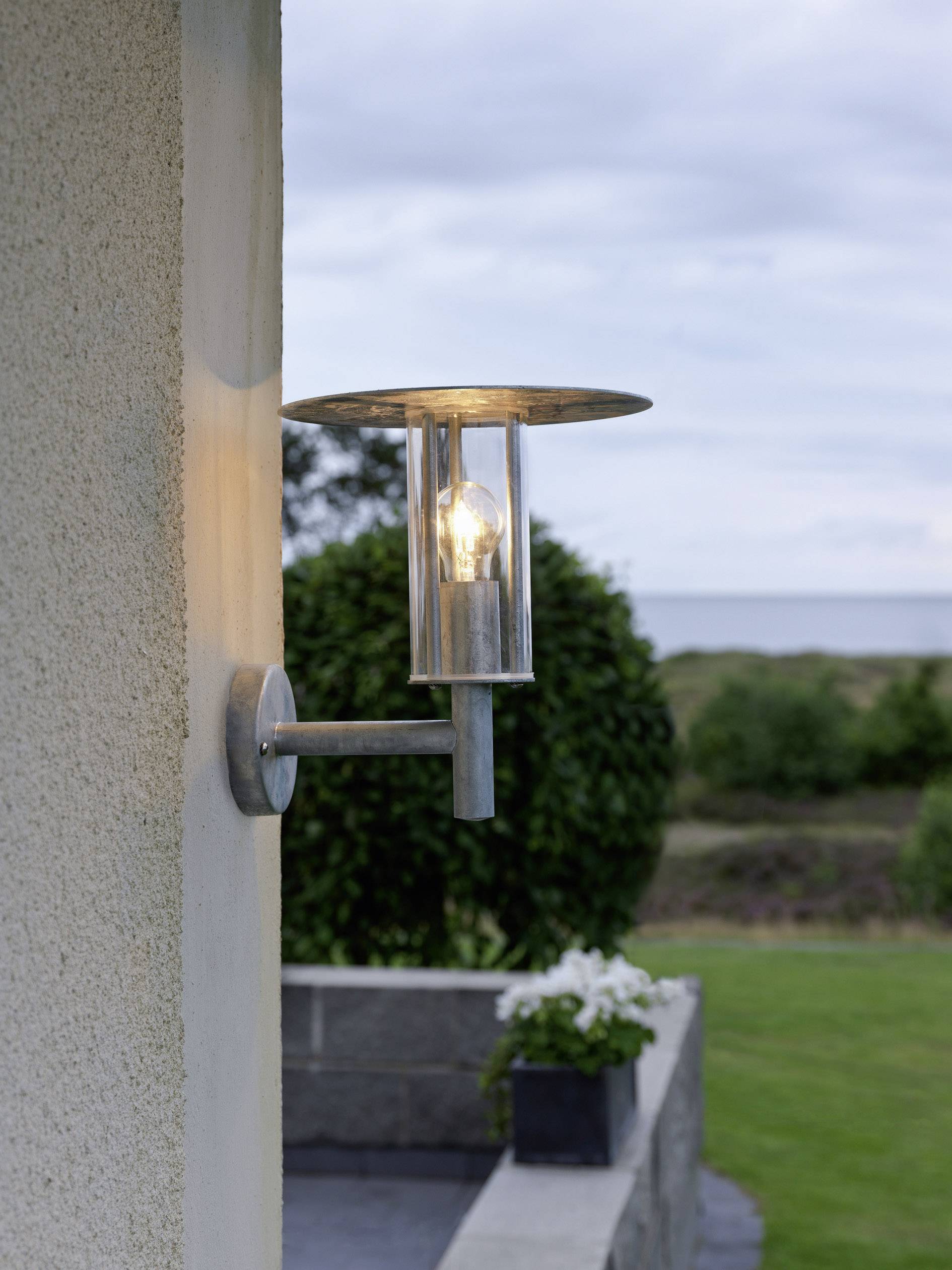 Außenbeleuchtung an einer Hauswand, mit einer brennenden Glühbirne in modernem Design, vor einem Garten mit Meerblick im Hintergrund.