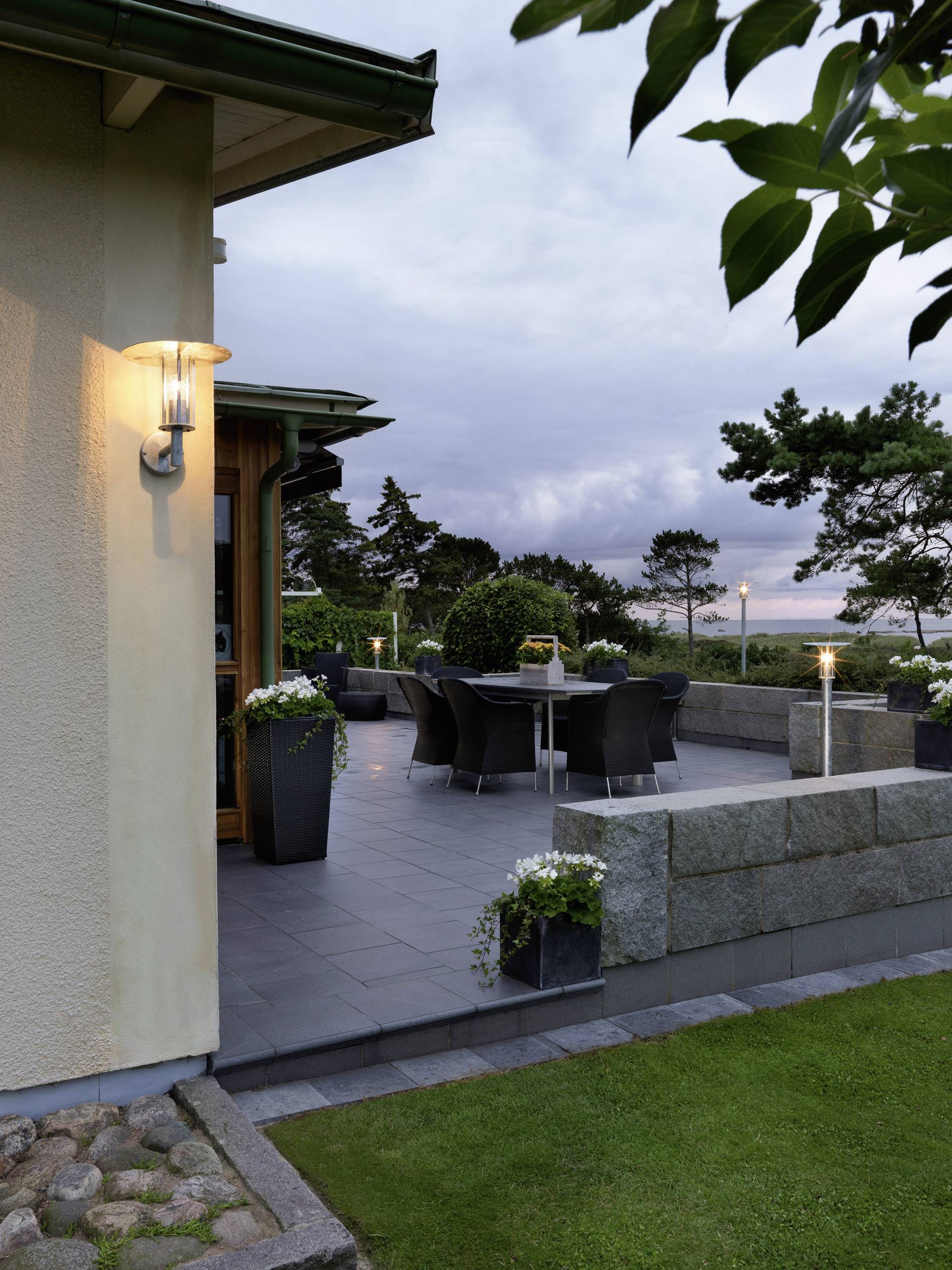 Terrasse eines Hauses bei Dämmerung, mit Tisch und Stühlen, von Bäumen umgeben, gedämpfte Beleuchtung schafft eine einladende Atmosphäre.
