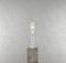 Eine moderne, transparente Stehlampe mit zylinderförmigem Glas-Schirm, montiert auf einem Betonblock, steht vor einer grauen Wand.