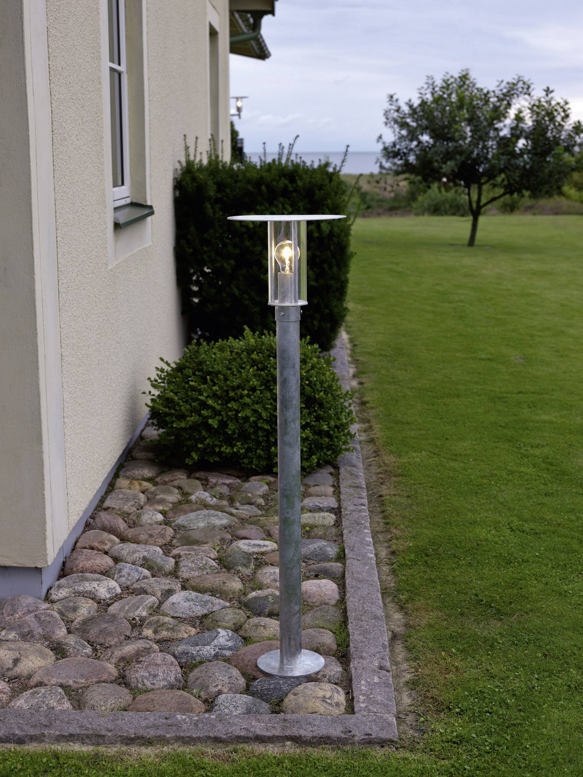 Gartenlampe neben einem Haus auf einem Kiesweg mit angrenzendem Rasen und Sträuchern. Im Hintergrund sind Bäume und ein bewölkter Himmel zu sehen.