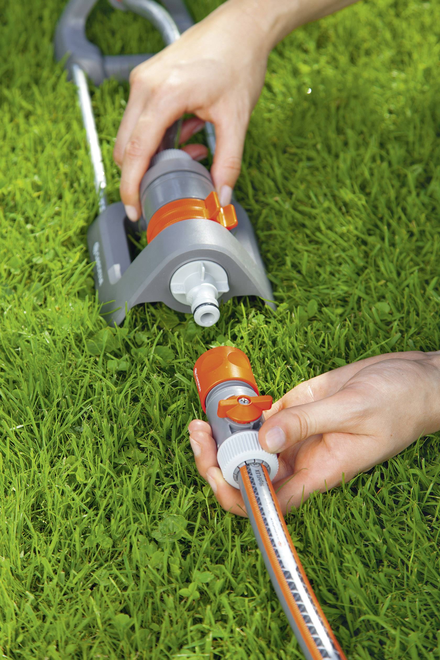 Zwei Hände verbinden einen Gartenschlauch mit einem Rasensprenger auf grünem Gras.