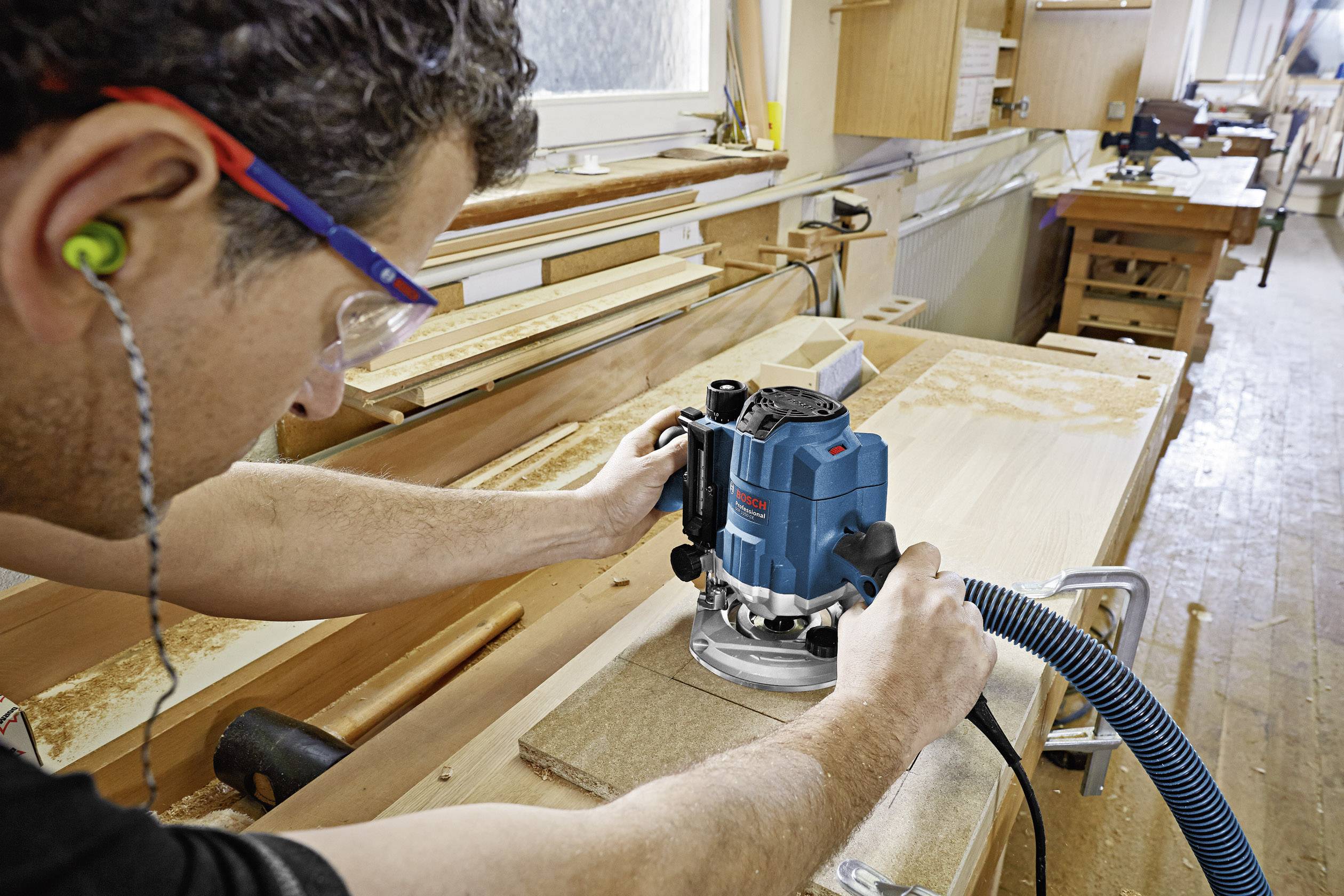 Ein Mann arbeitet in einer Werkstatt mit einer blauen Fräsmaschine auf einem Holzbrett, trägt Sicherheitsbrille und Ohrstöpsel.