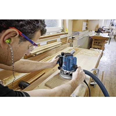 Ein Mann arbeitet in einer Werkstatt mit einer blauen Fräsmaschine auf einem Holzbrett, trägt Sicherheitsbrille und Ohrstöpsel.