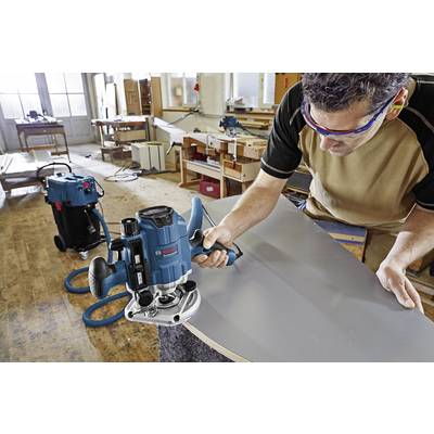 Ein Handwerker bearbeitet mit einer Elektrowerkzeugmaschine eine große Holzplatte in einer Werkstatt. Im Hintergrund sind weitere Werkzeuge und Werkbänke zu sehen.