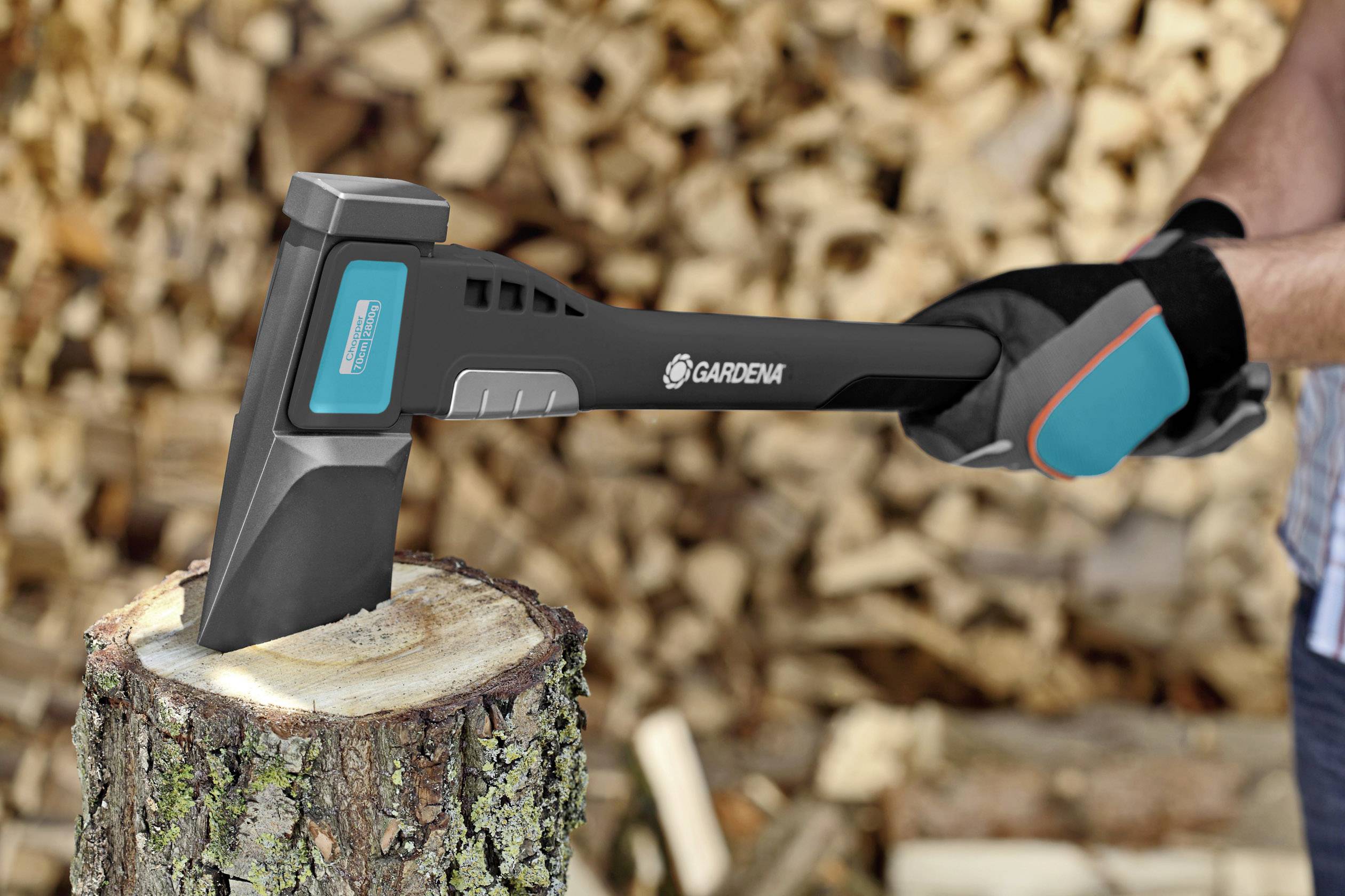 Person hackt Holz mit einer Axt. Im Hintergrund ein Stapel gehacktes Feuerholz. Die Szene zeigt eine Outdoor-Arbeitssituation.