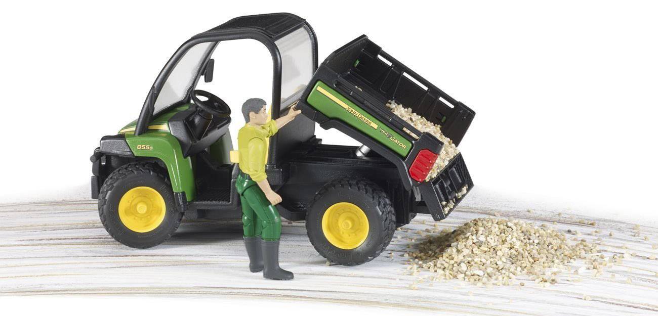 Ein Modelltraktor mit Anhänger kippt Sand ab. Eine Spielzeugfigur in grüner Hose interagiert mit dem Anhänger.