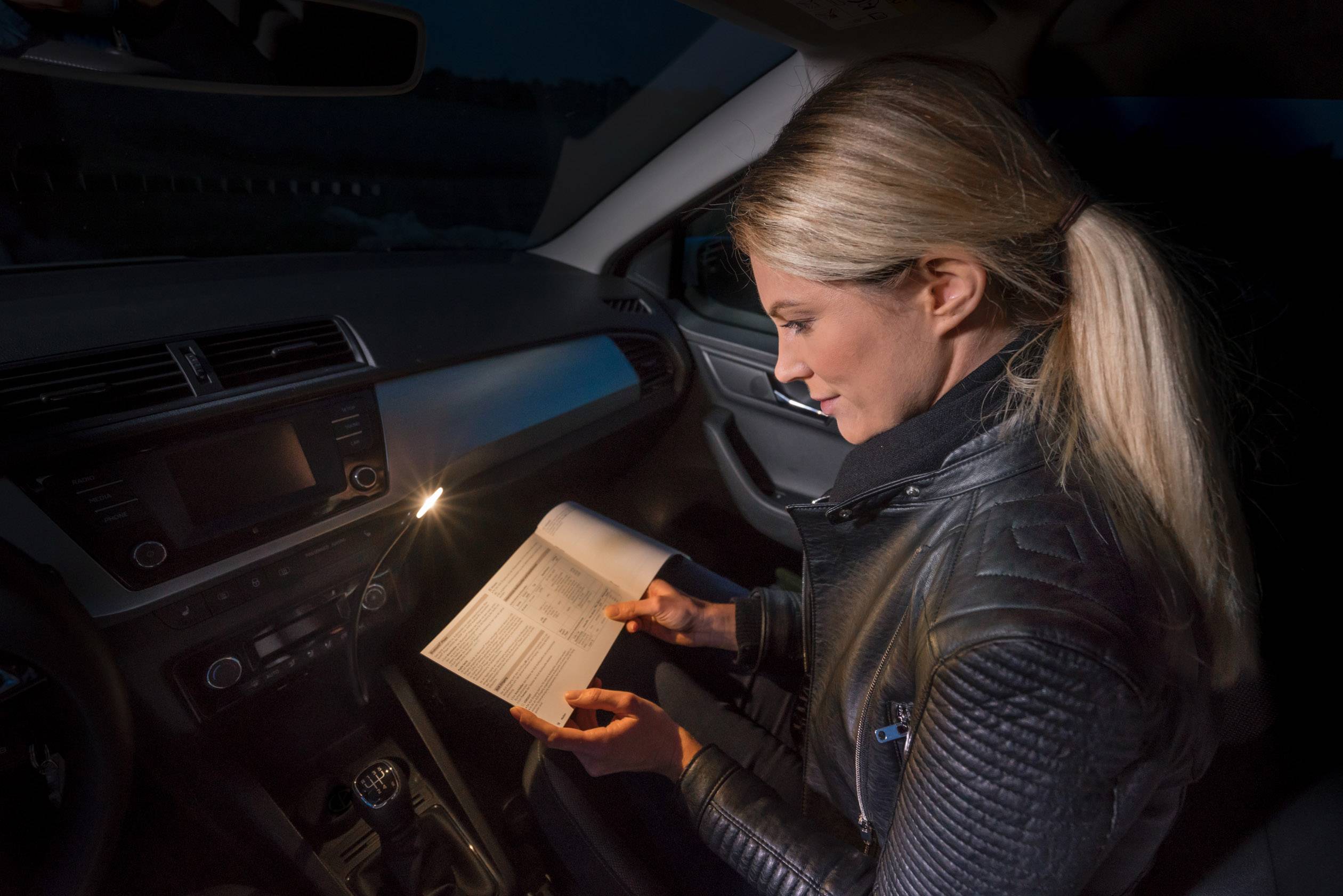 Eine Frau sitzt im Auto bei Dunkelheit und liest ein Handbuch. Sie beleuchtet die Seite mit einem kleinen Licht, das im Auto montiert ist.