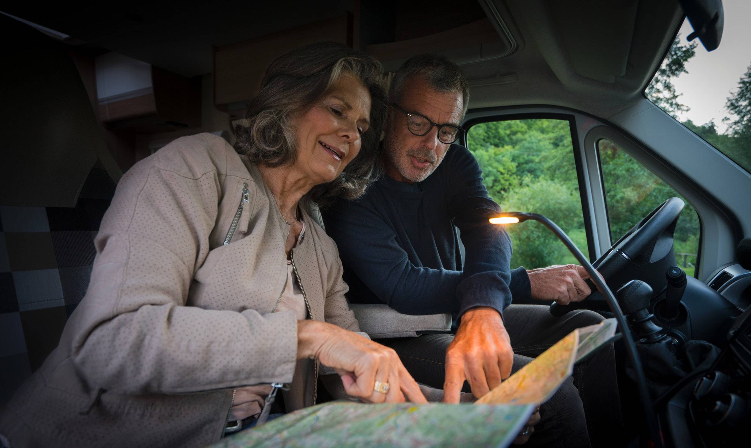 Ein älteres Paar sitzt im Auto und betrachtet eine Karte. Sie scheinen eine Route zu planen, während sie von einer Lampe beleuchtet werden.