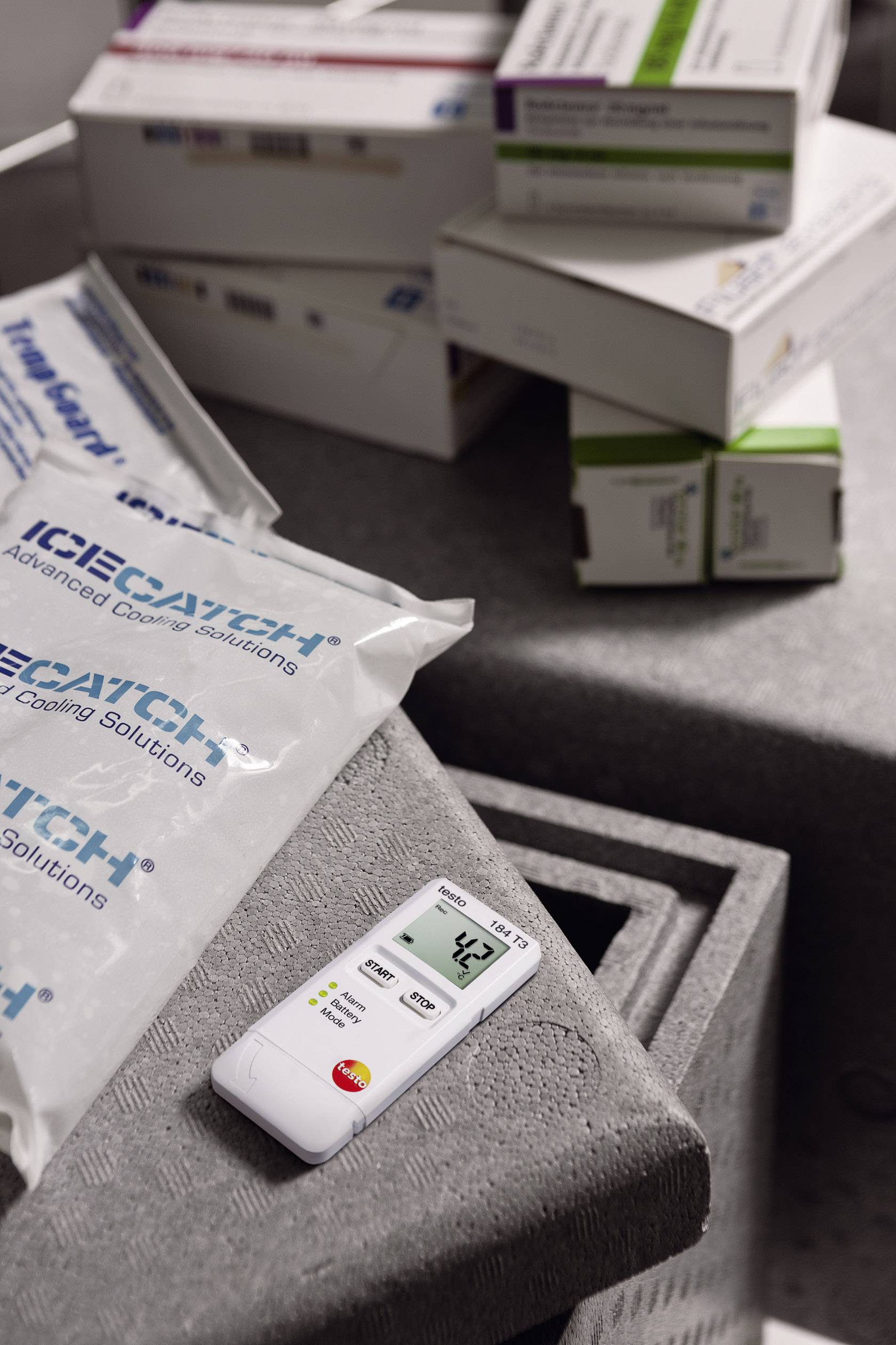 Ein digitales Thermometer zeigt 4,3 Grad Celsius an und liegt auf einer Isolierbox mit Kühlpackungen und Medikamentenkartons im Hintergrund.