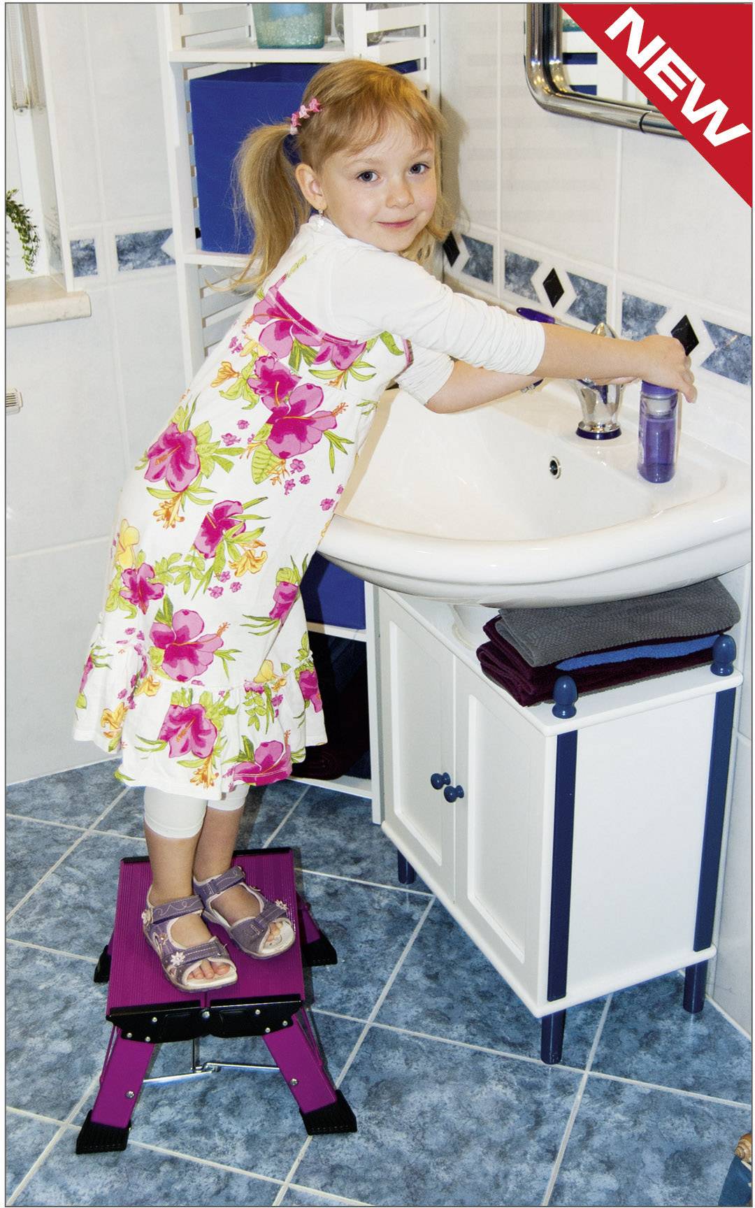Ein lächelndes Kind steht auf einem Hocker vor einem Waschbecken und wäscht die Hände. Es trägt ein Kleid mit Blumenmuster.