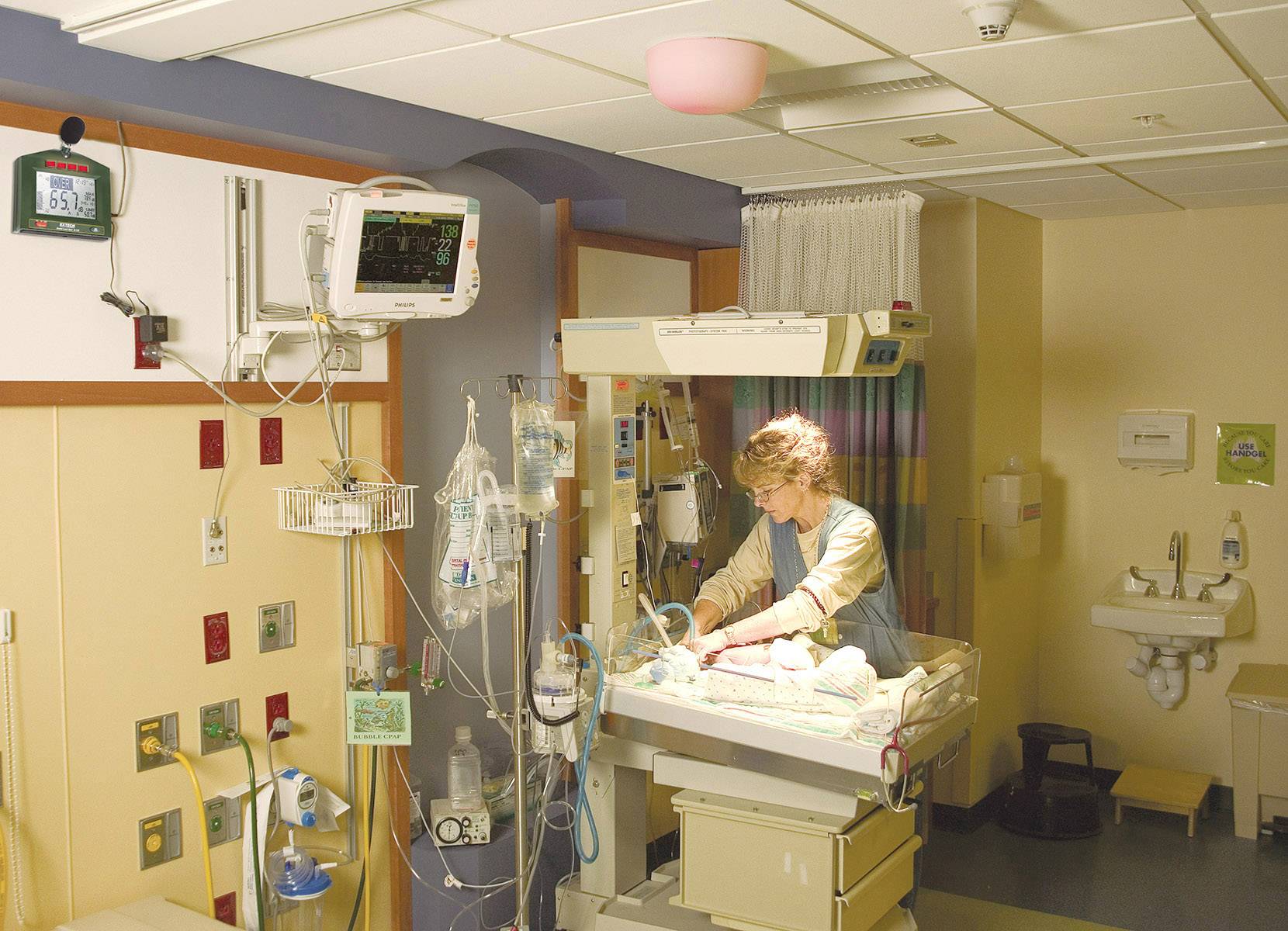 Eine Krankenschwester kümmert sich um ein Neugeborenes in einem Krankenhauszimmer, das mit medizinischen Geräten ausgestattet ist.