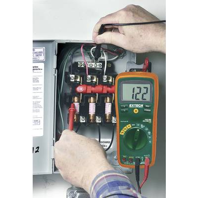 Ein Elektriker misst die Spannung mit einem Multimeter an einem geöffneten Sicherungskasten. Das Display zeigt '122'.