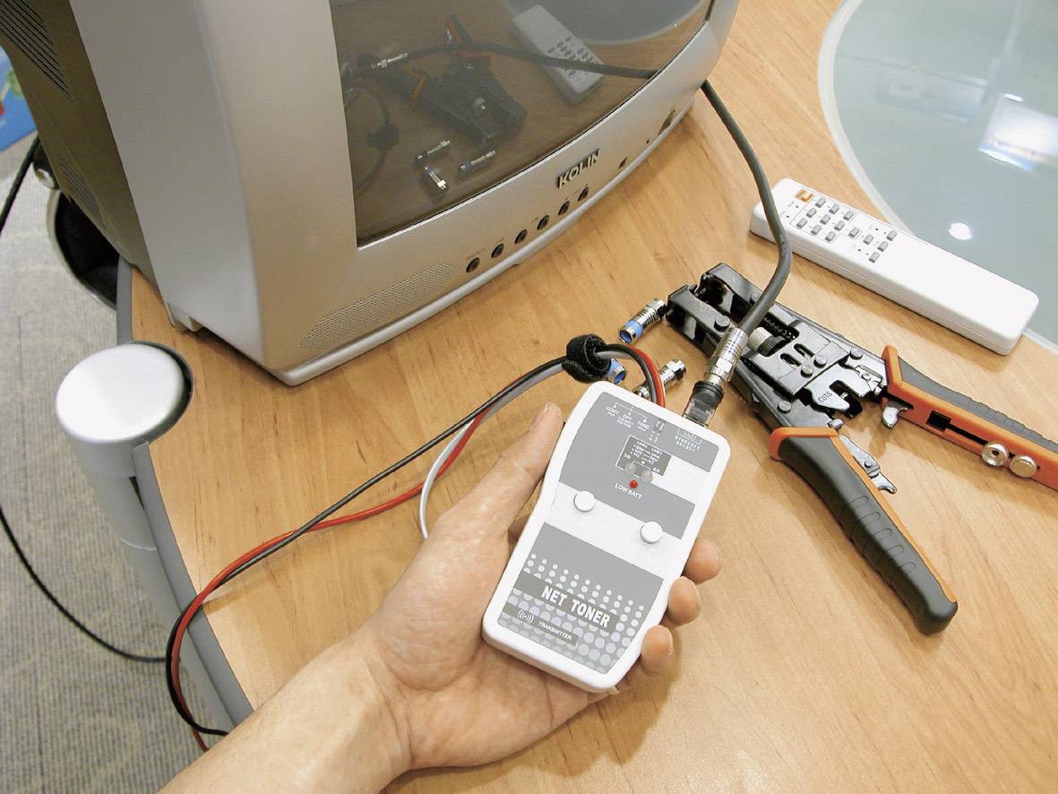 Eine Hand hält ein Netzwerkkabeltester-Gerät neben einem alten Fernseher. Werkzeuge und Kabel sind auf einem Holztisch verteilt.