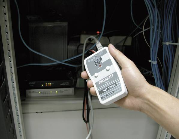 Das Bild zeigt einen handgehaltenen, grauen VOLTCRAFT Net Toner Leitungssucher vor einem Serverschrank mit Netzwerkkabeln und Hardware. Das rechteckige Testgerät hat eine graue Oberfläche mit verschiedenen Anzeigen und Kontrollleuchten zur Identifikation von Leitungen. Im Hintergrund sind mehrere Netzwerkgeräte und viele blaue und graue Kabel zu sehen, die durch das Gehäuse verlaufen.