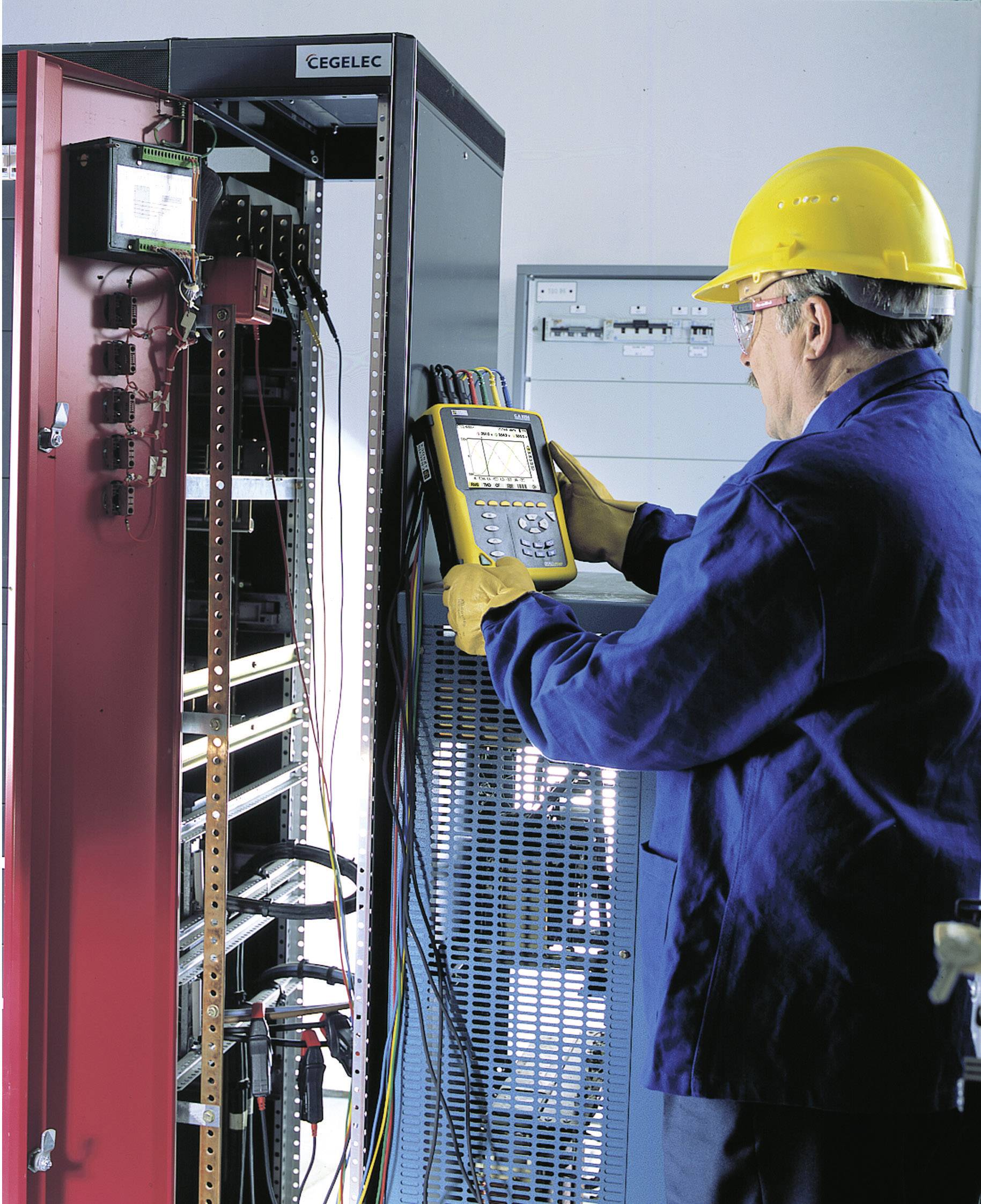 Ein Techniker mit gelbem Schutzhelm prüft elektrische Schaltanlagen mit einem Handmessgerät in einem Industriebetrieb.