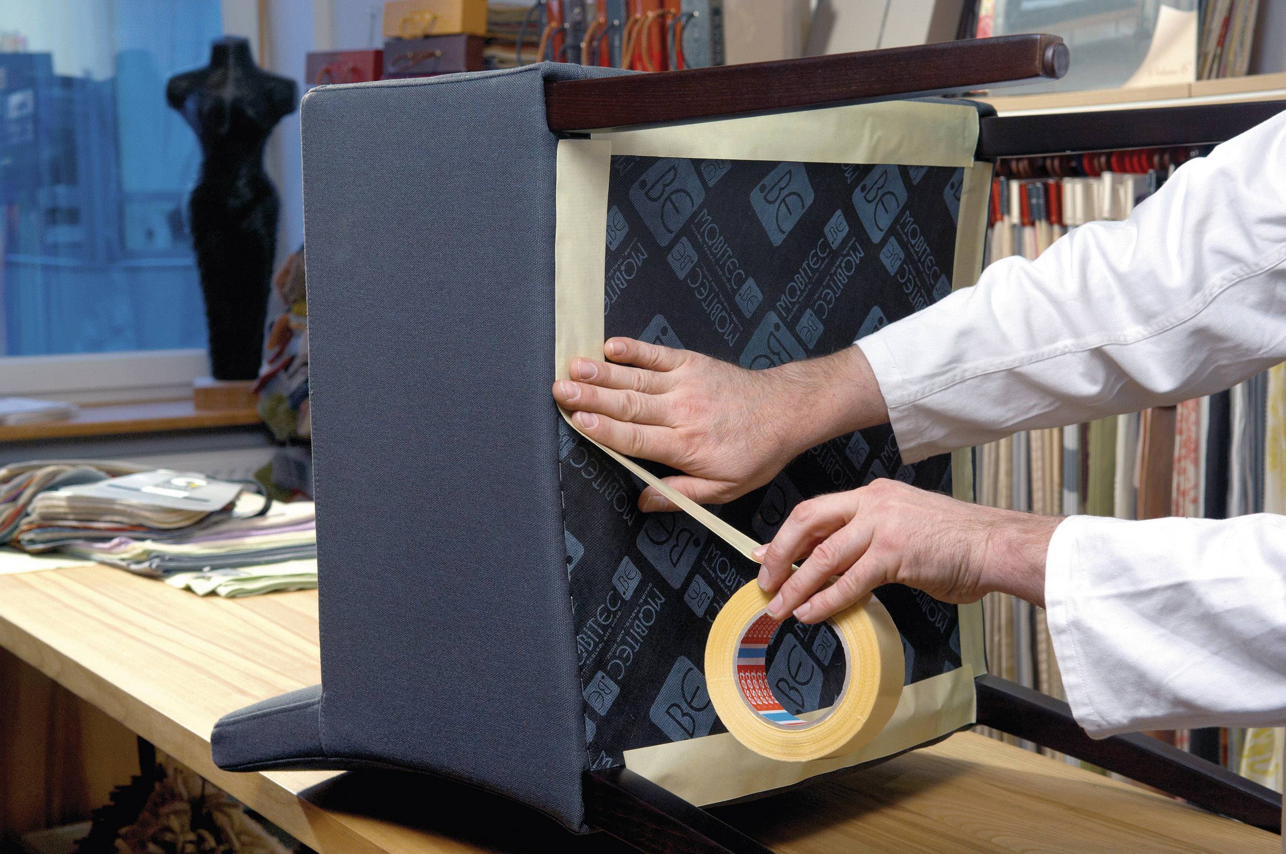 Eine Person in einem weißen Kittel befestigt Klebeband auf der Rückseite eines Sessels. Im Hintergrund sind Stoffmuster zu sehen.