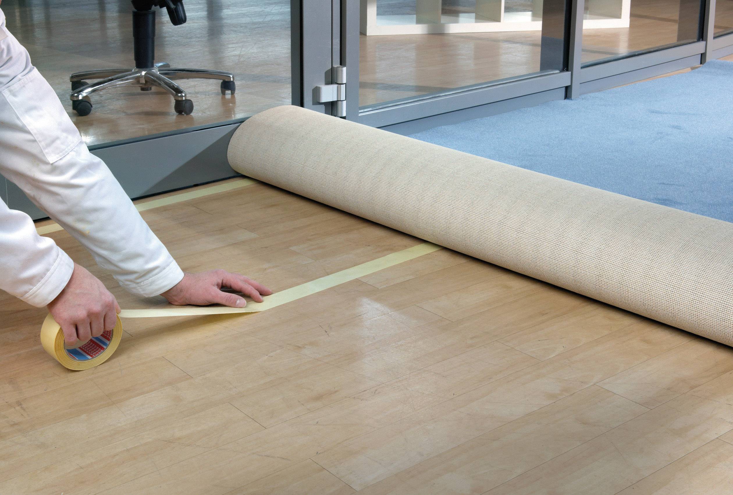 Eine Person klebt Klebeband auf einen Holzboden, um einen Teppich an der Übergangsstelle zu fixieren. Im Hintergrund sind Büromöbel sichtbar.