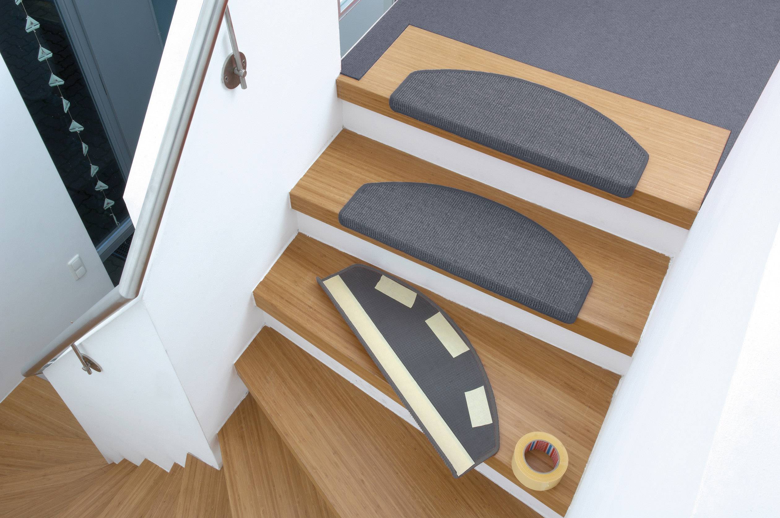 Treppenstufen mit Antirutschmatten aus Teppichmaterial, eine lagert am Rand. Daneben ein Klebeband, vermutlich zur Befestigung.