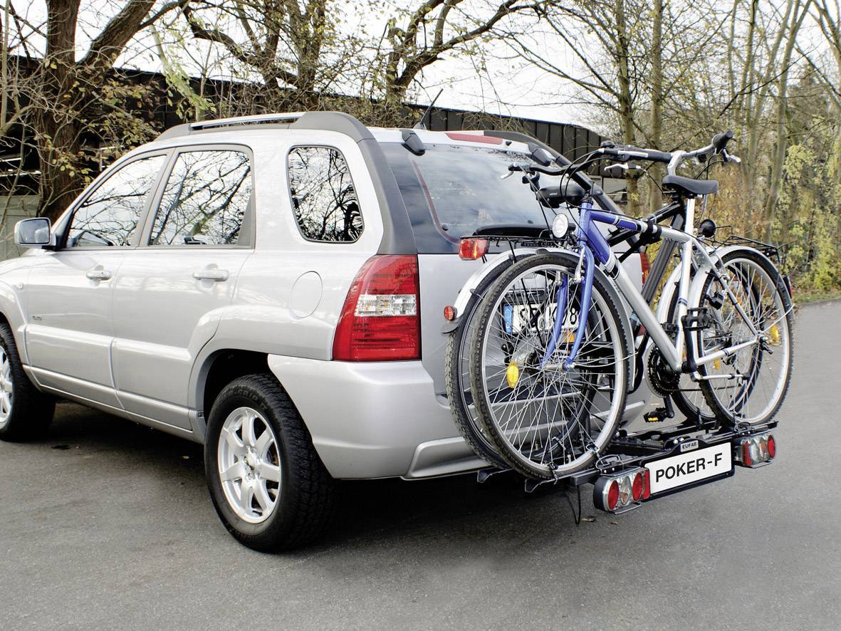 Ein silberner SUV mit zwei Fahrrädern auf einem Fahrradträger am Heck, geparkt auf einer Straße mit Bäumen im Hintergrund.