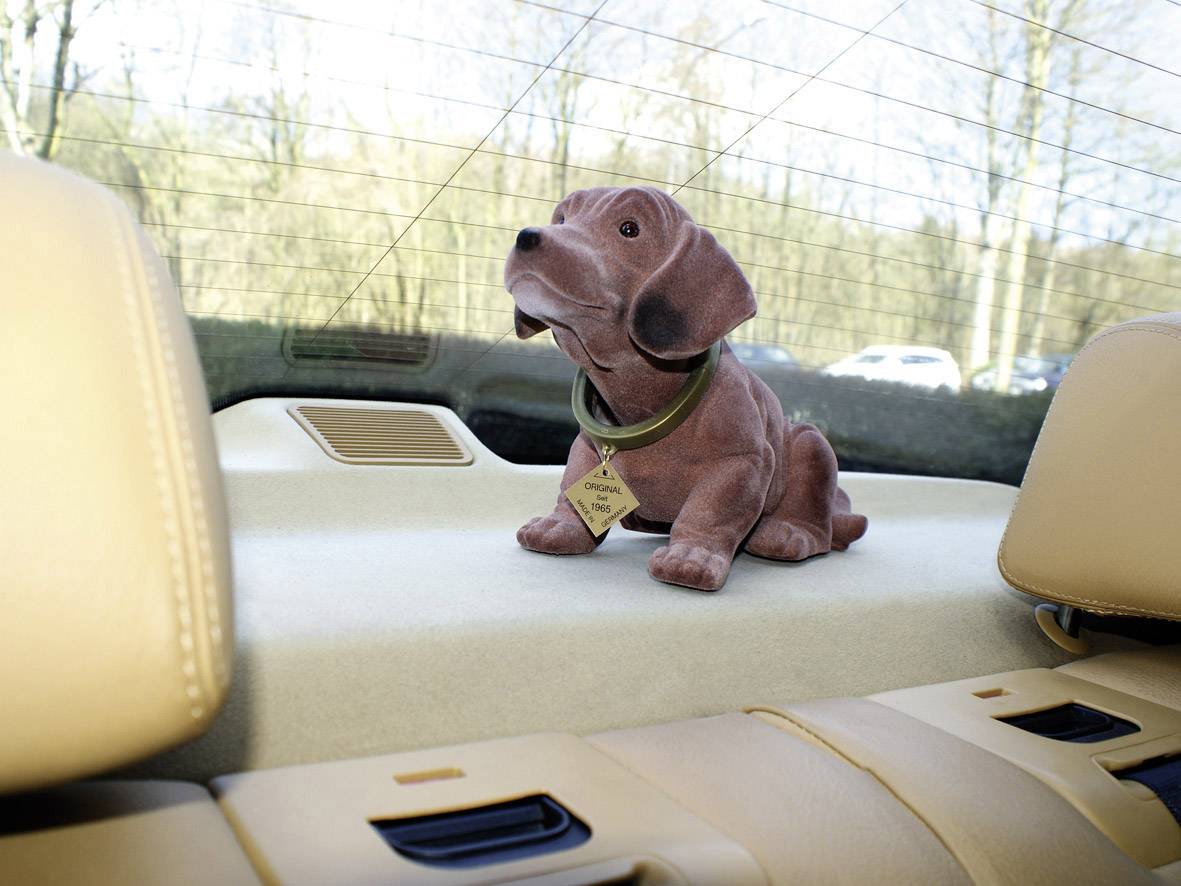 Ein Wackeldackel in einem Auto steht auf der Hutablage hinter den Rücksitzen. Der kleine Hund hat ein braunes Stofffell und trägt ein Halsband.