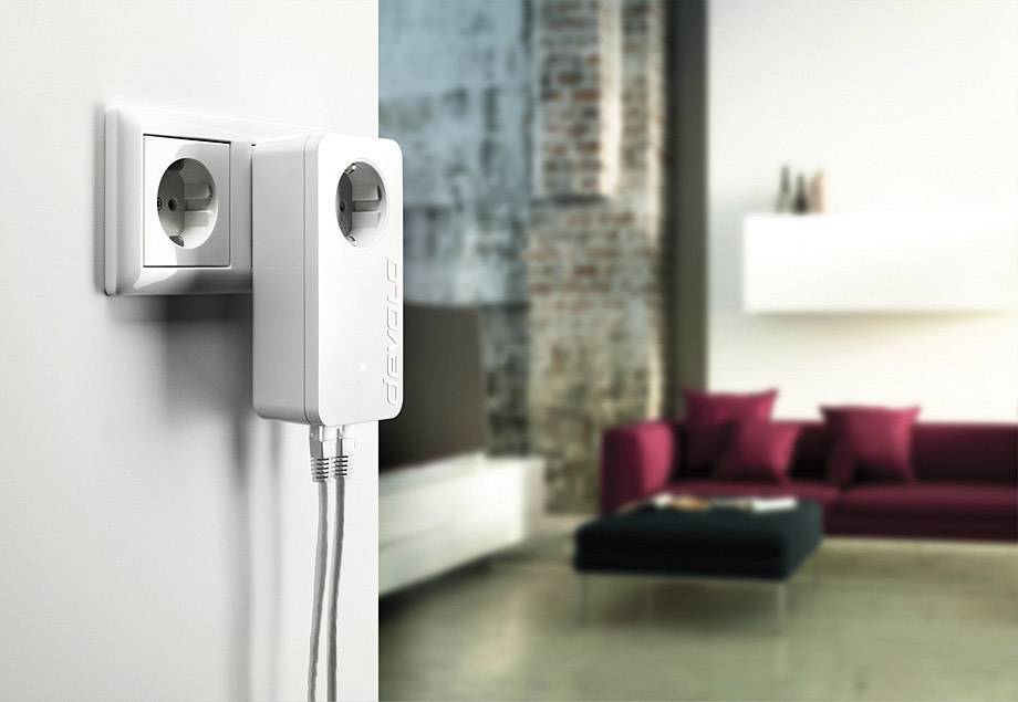 Ein weißer Adapter ist in eine Wandsteckdose eingesteckt, mit zwei Kabeln, die darunter verlaufen. Im Hintergrund ein Wohnzimmer mit Sofa.