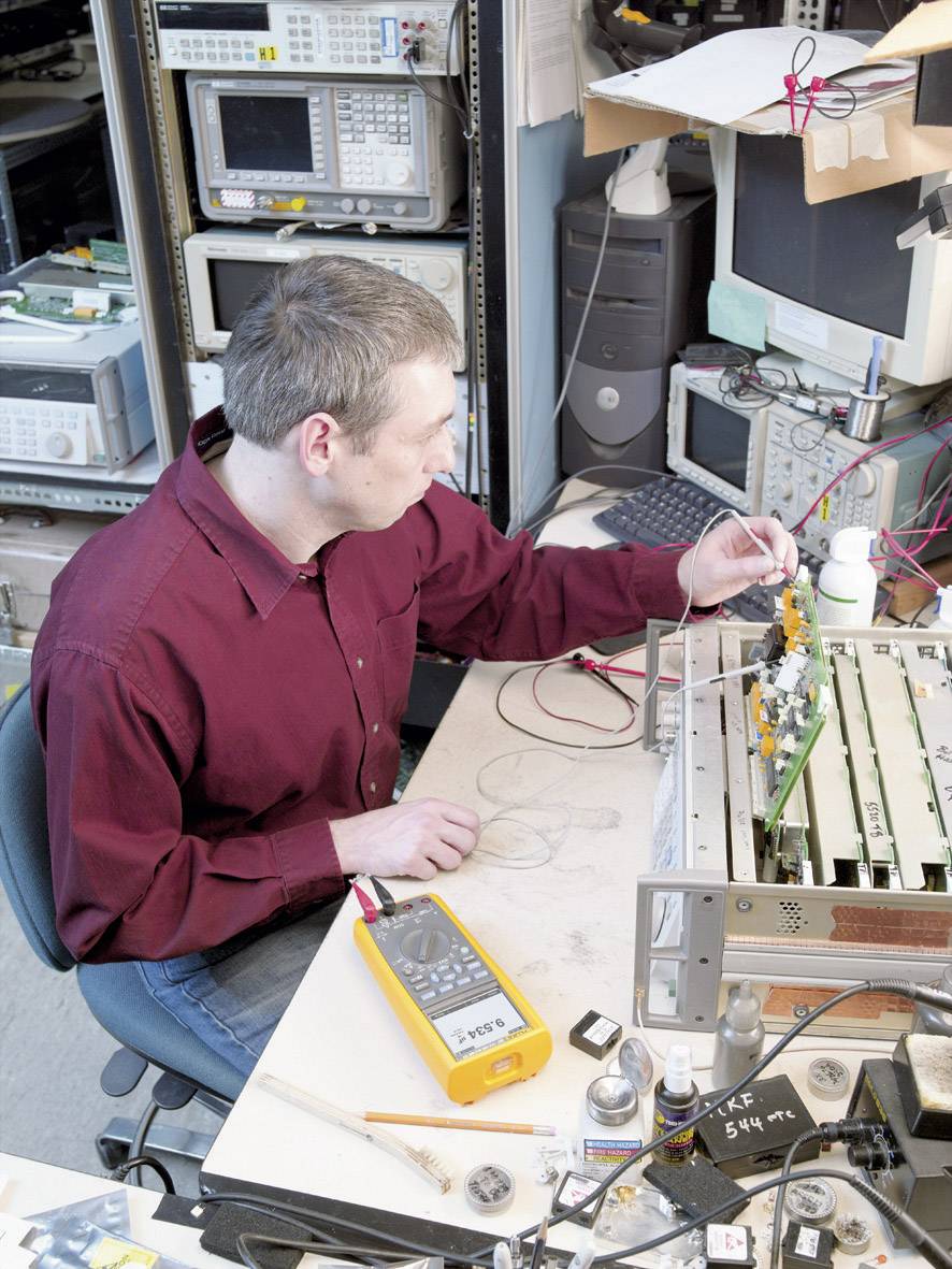 Eine Person in einem technischen Labor arbeitet an einer Leiterplatte mit elektronischen Messgeräten und Werkzeugen auf dem Tisch.