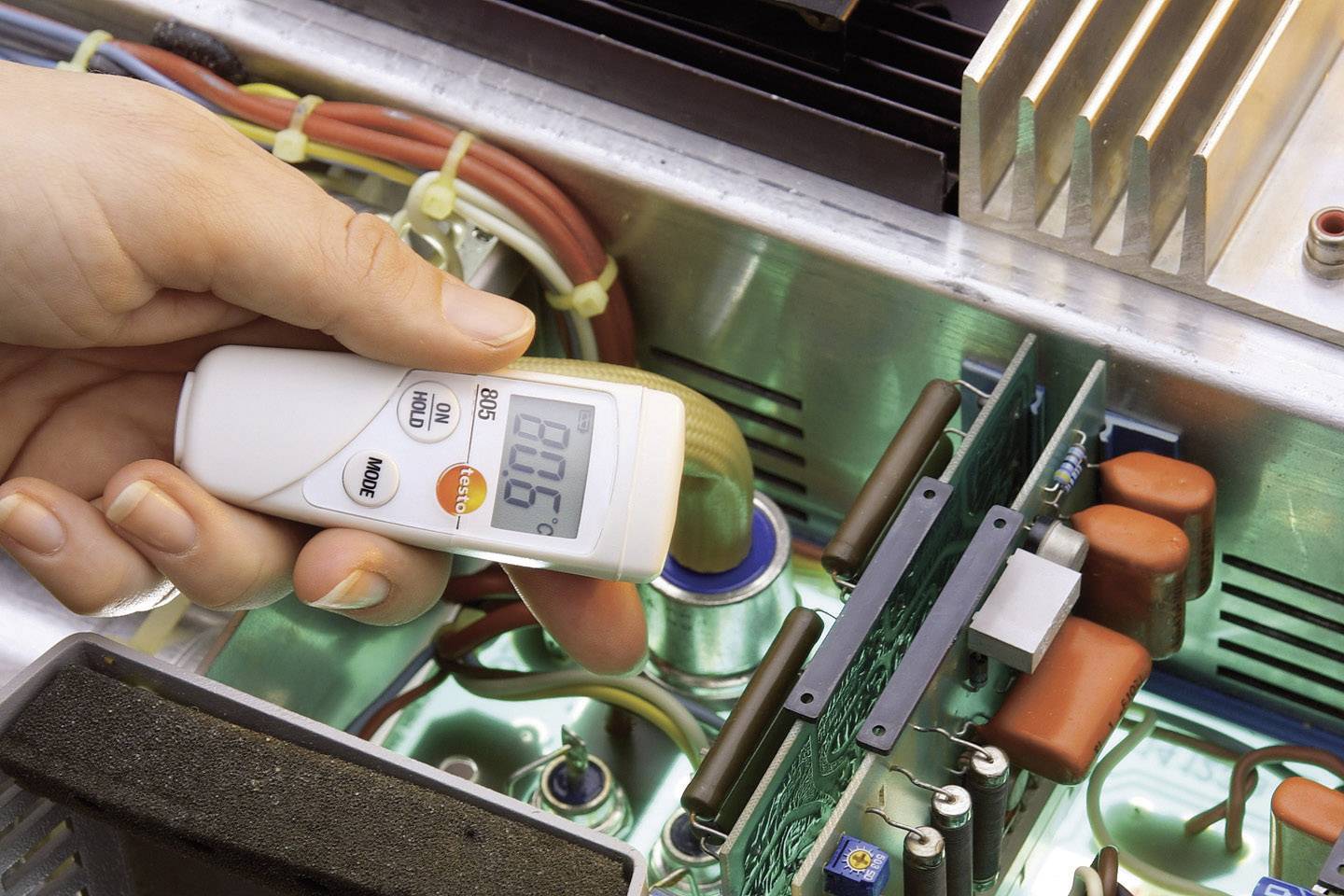 Eine Person misst die Temperatur in einem elektronischen Gerät mit einem Infrarot-Thermometer. Das Display zeigt '90.8'.