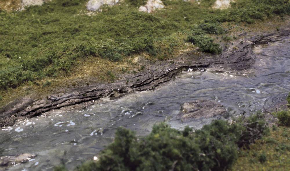 Ein kleiner Fluss fließt durch eine grüne, grasbewachsene Landschaft. Steine sind am Ufer sichtbar.